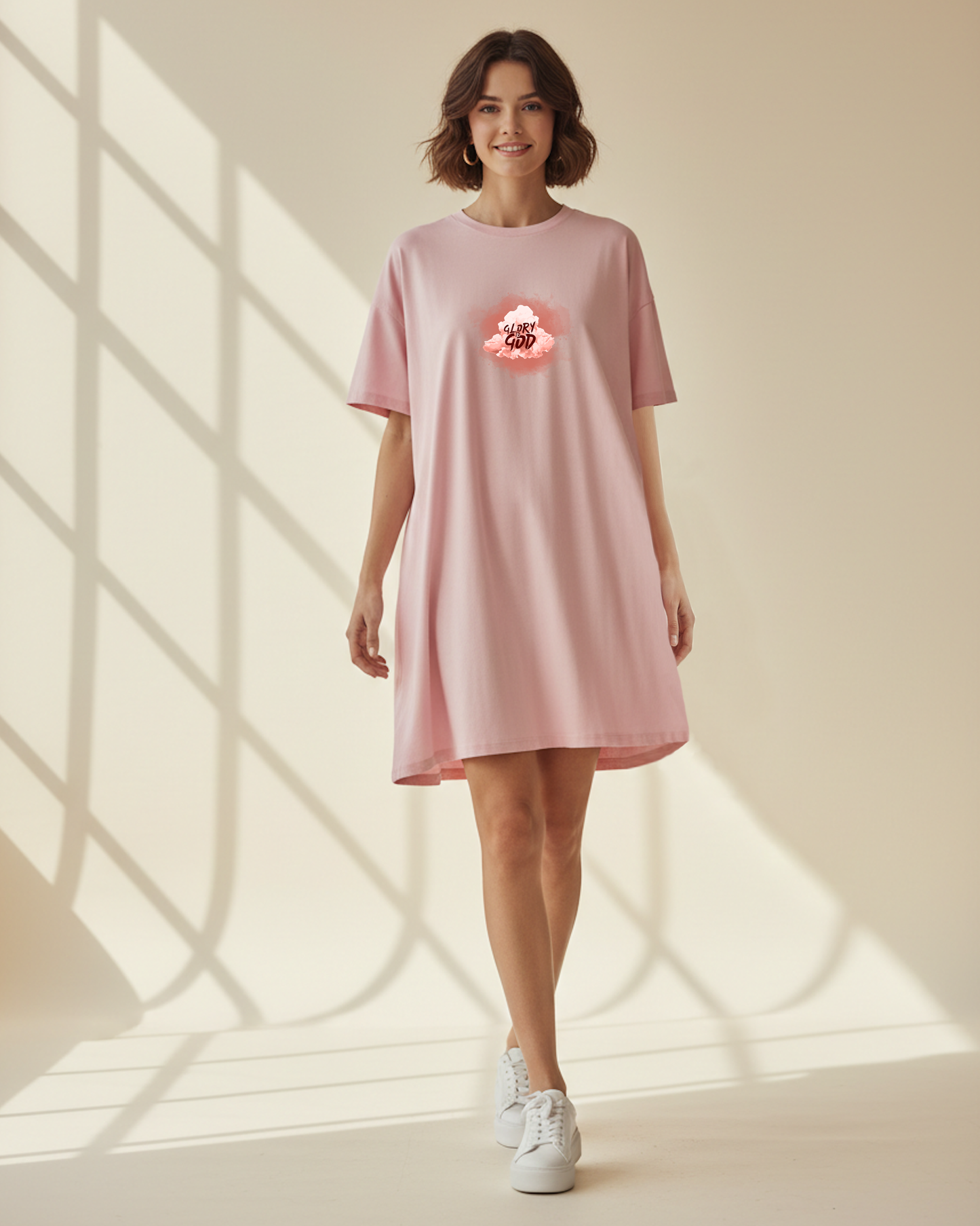 Woman wearing a pink t-shirt dress with a graphic Glory to God text in a cloud design in a bright room with sunlight filtering through.