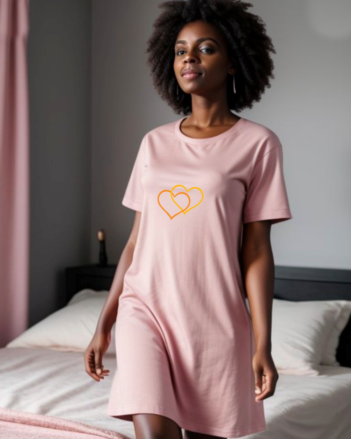 A woman stands in a bedroom, dressed in a pink t-shirt dress featuring the Heavenly Dreamwear logo of two intertwined heart shapes printed on the front.