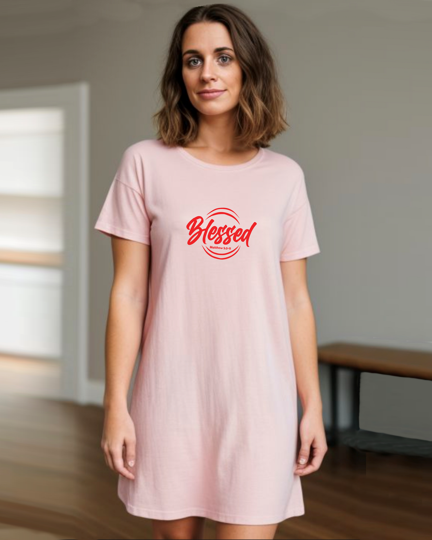 A woman in a room wearing a pink Heavenly Dreamwear t-shirt dress with a red logo comprised of the text "Blessed" on top of a broken double circle with a biblical scripture reference beneath the text.