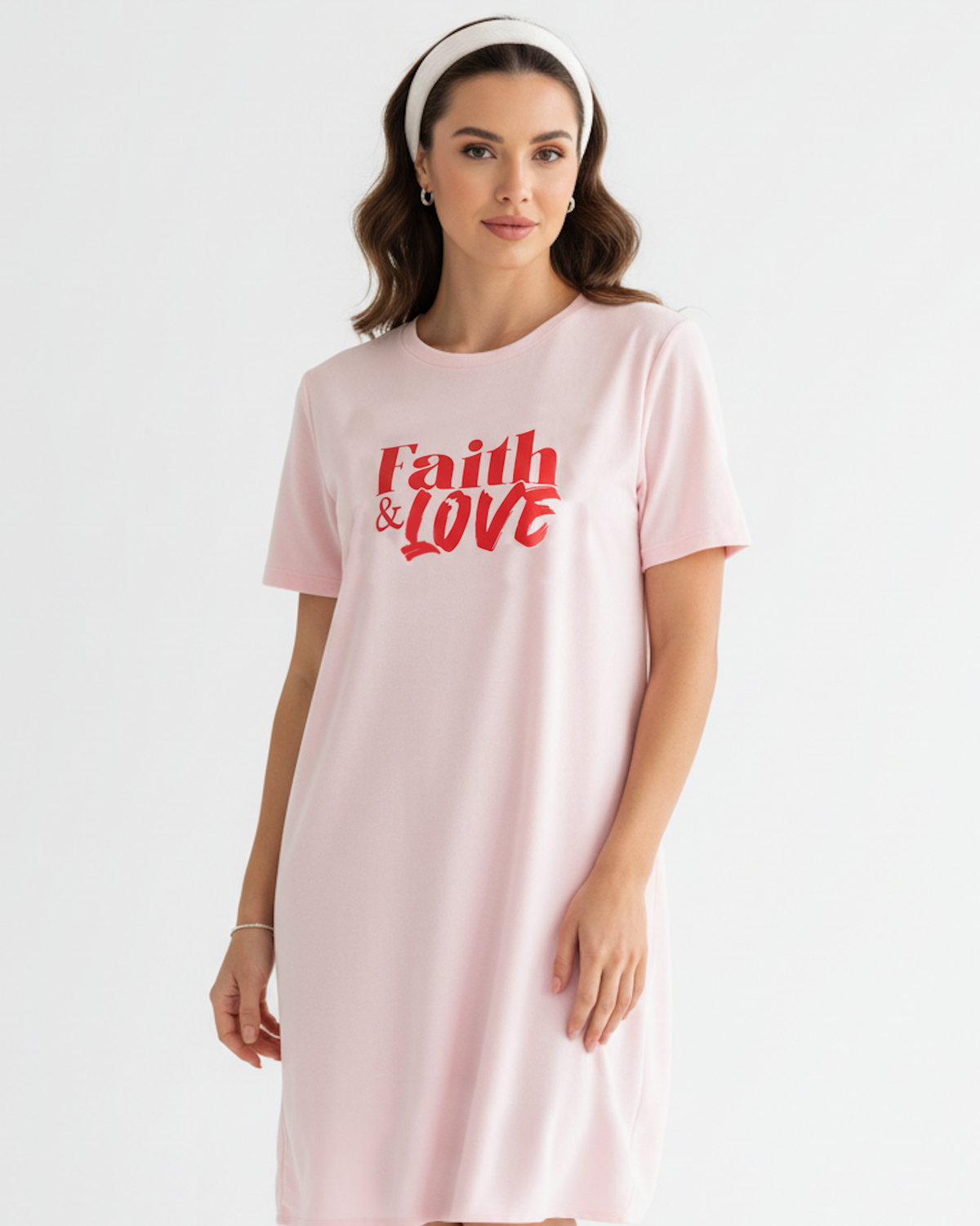 A woman wearing a light pink short-sleeve t-shirt dress with 'faith & LOVE' printed in red across the chest, paired with a white headband, against a plain white studio background.