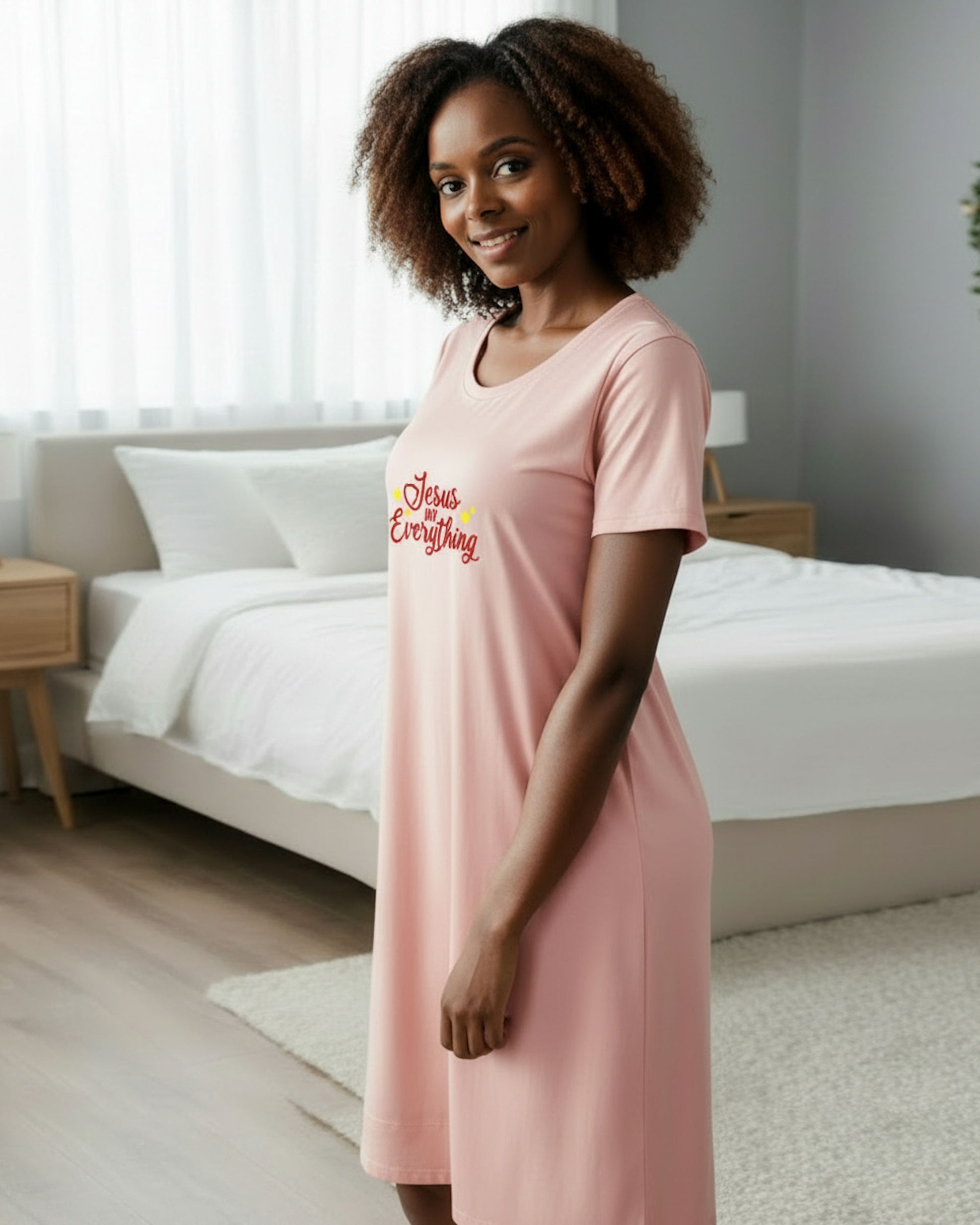A woman wearing a light pink short-sleeve nightgown with the phrase 'Jesus MY Everything' printed in red and yellow on the chest, standing in a modern bedroom with a white bed in the background.