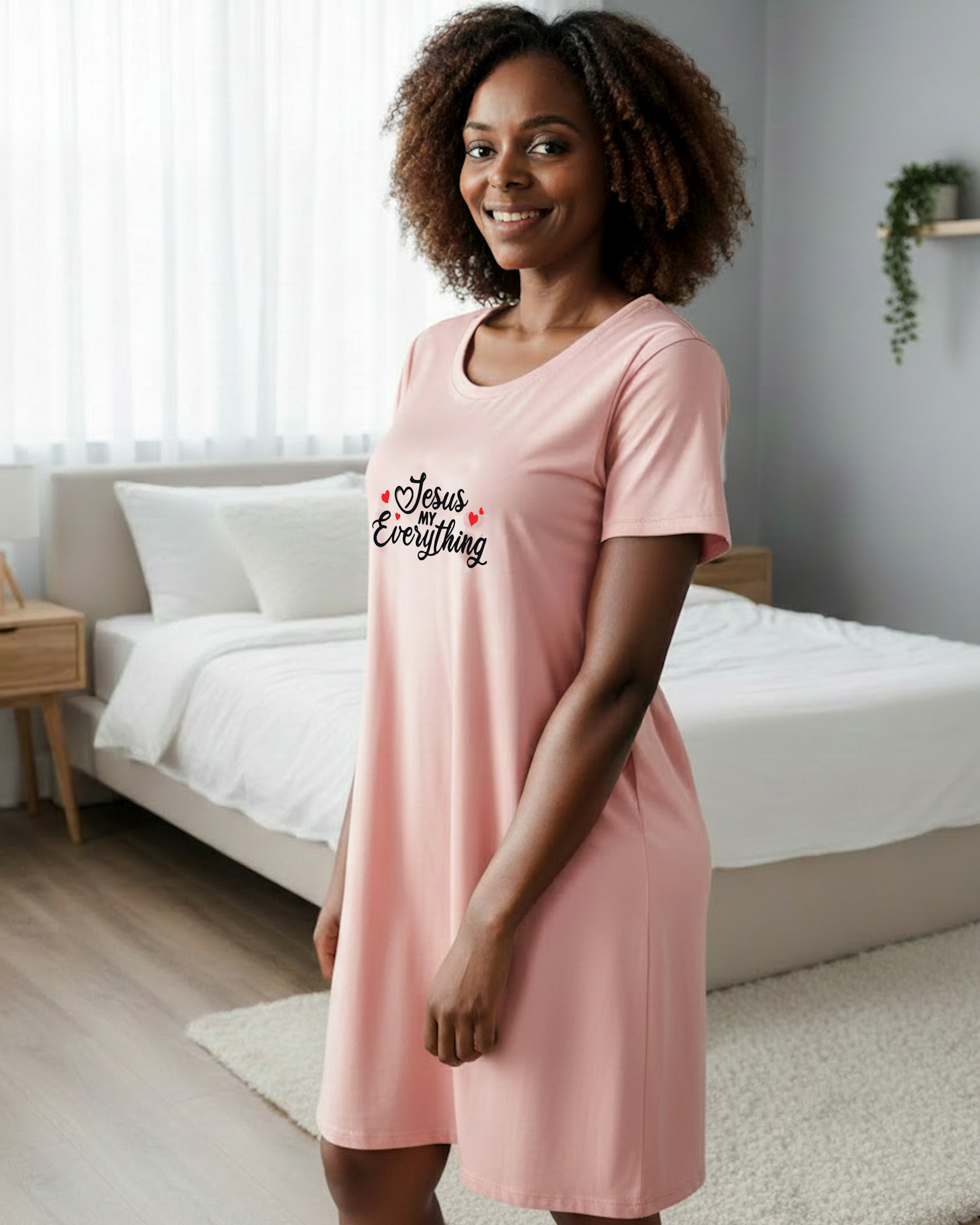 Woman wearing a light pink t-shirt dress with  black 'Jesus MY Everything' text and 4 hearts logo in a bedroom setting
