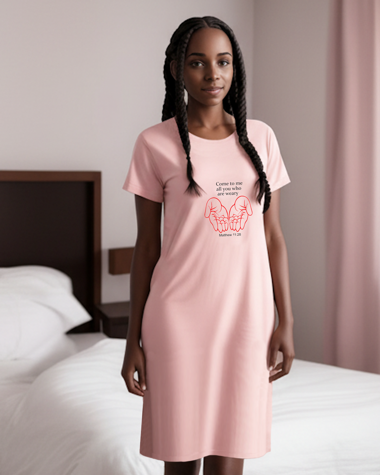 A woman with long braids stands in a bedroom wearing a light pink short-sleeve t-shirt dress with a red graphic of two hands holding hearts with scripture 'Come to me all who are weary' from Matthew 11:28.