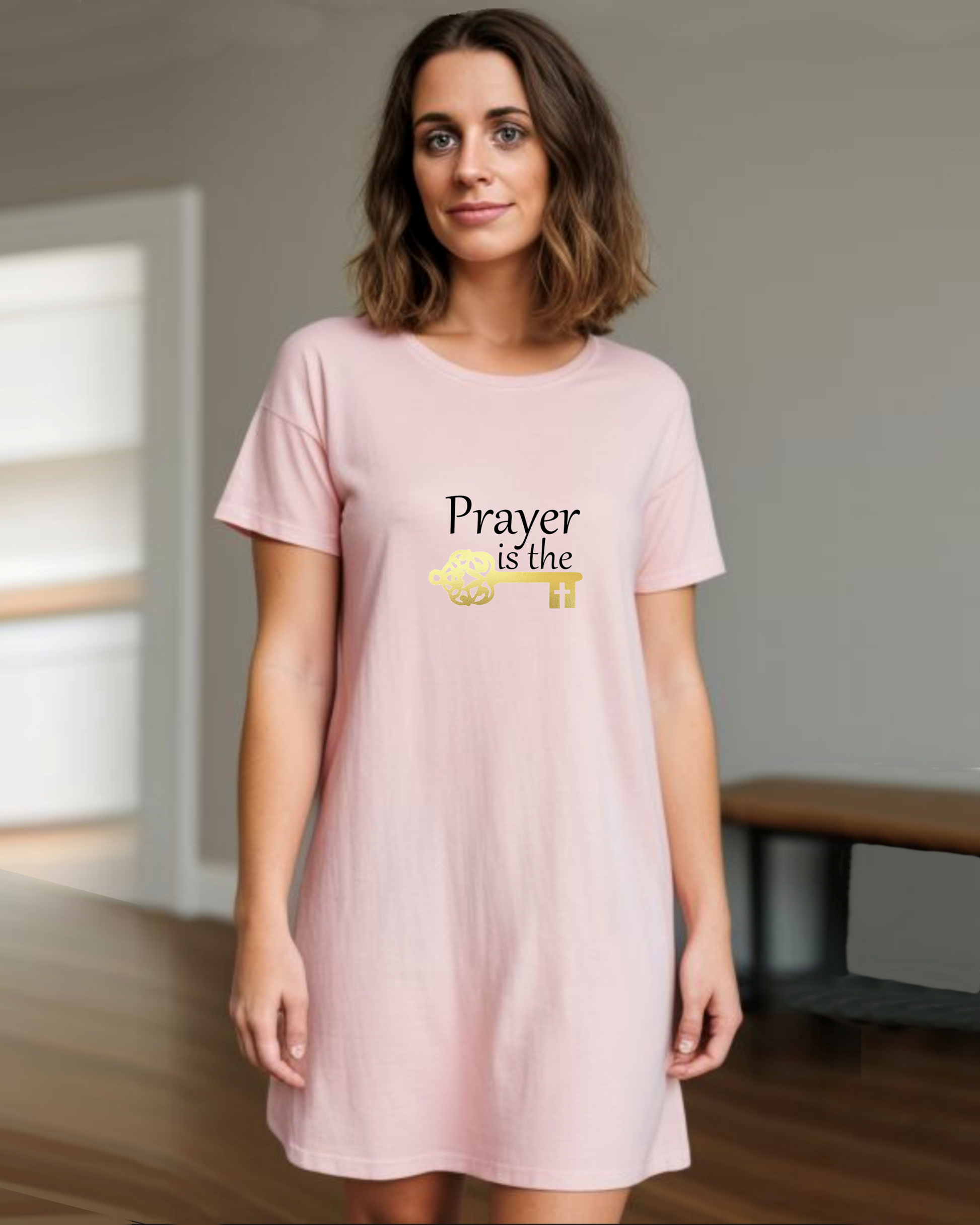 A woman is wearing a pink cotton t-shirt dress featuring a design by Heavenly Dreamwear.  The design includes black text that reads, "Prayer is the," followed by a gold key symbol.