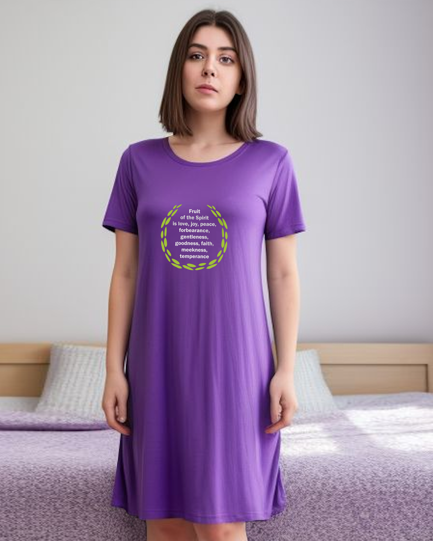 A woman in a bedroom wearing a purple t-shirt dress with a logo stating the fruit of the Holy Spirit in white text surrounded by green leaves.