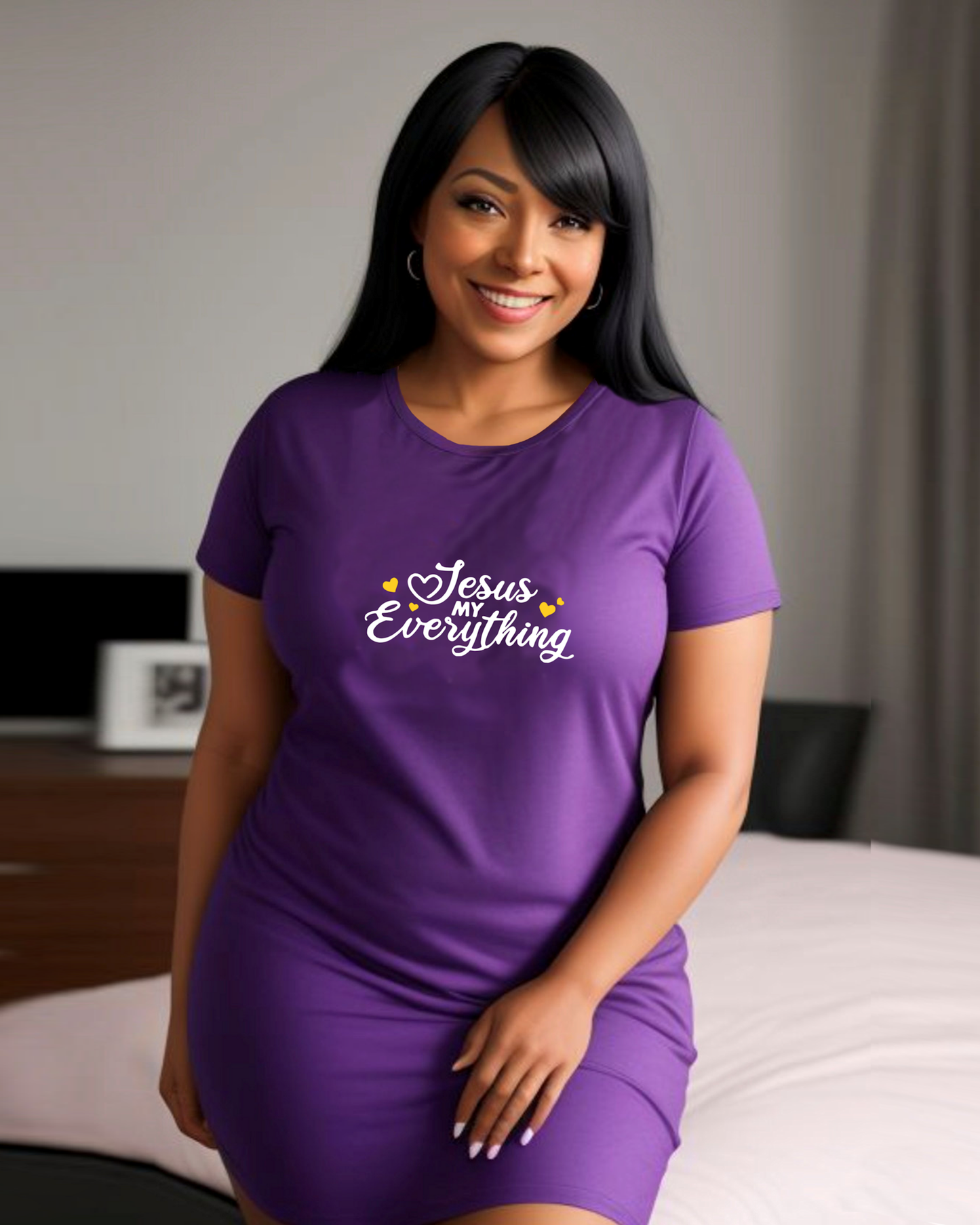 A woman in a bedroom wearing a purple Heavenly Dreamwear dress with a logo made up of white text, "Jesus MY Everything", with yellow hearts beside it, printed on the front.