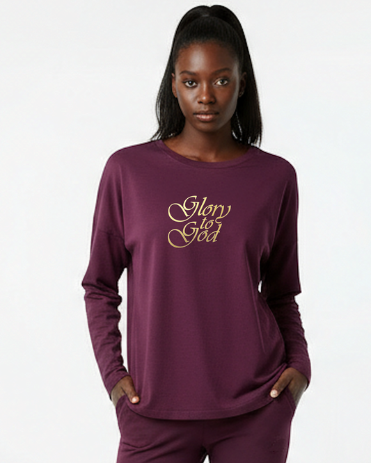 A woman modeling a long-sleeve crewneck top in deep plum purple with 'Glory to God' printed in gold cursive across the chest, paired with matching purple pants against a plain white background.