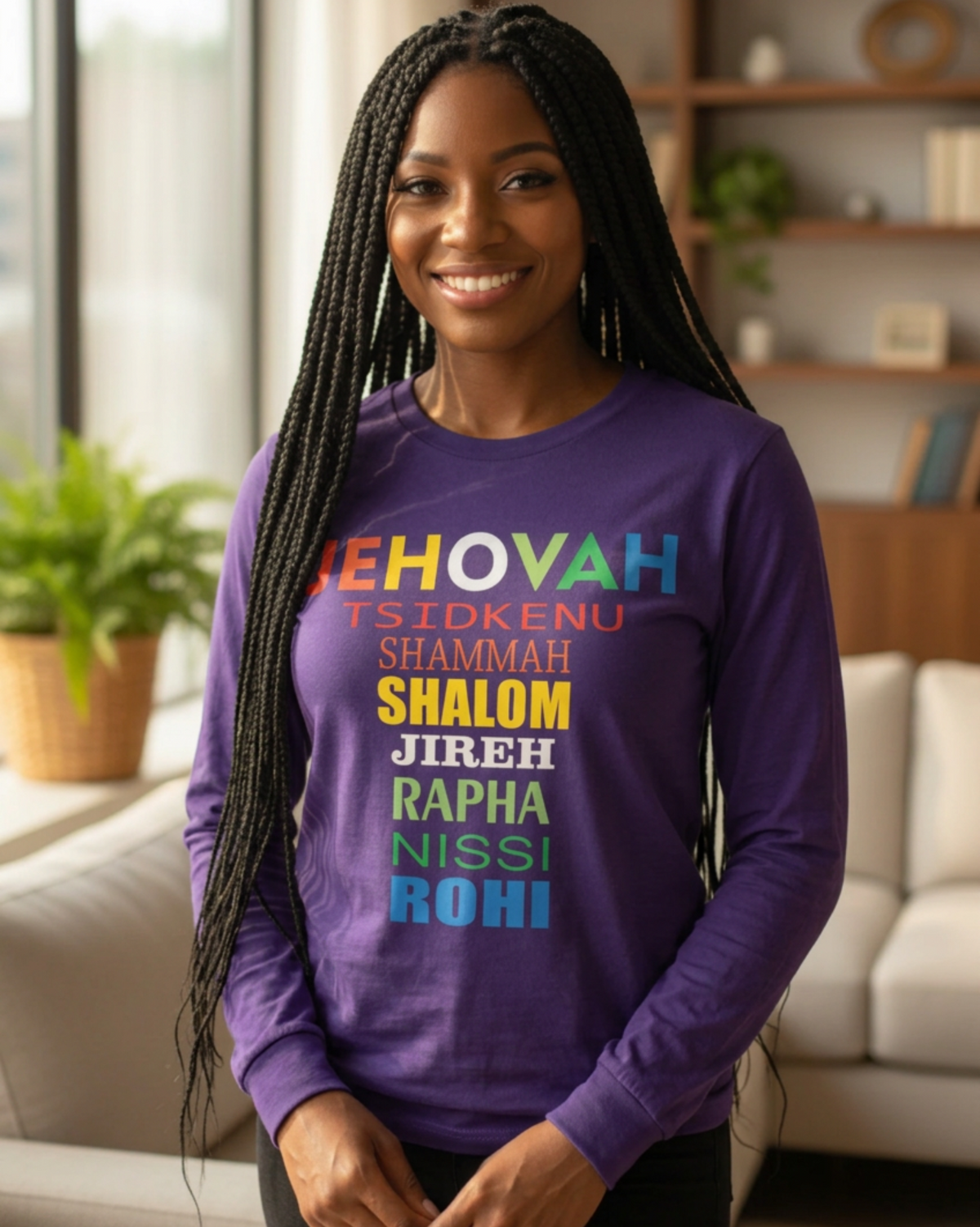 A smiling woman with long black braids stands indoors wearing a purple long-sleeve shirt with colorful Hebrew and English text on the front, paired with dark pants. The background shows a softly blurred living room with a sofa, plants, and shelves.