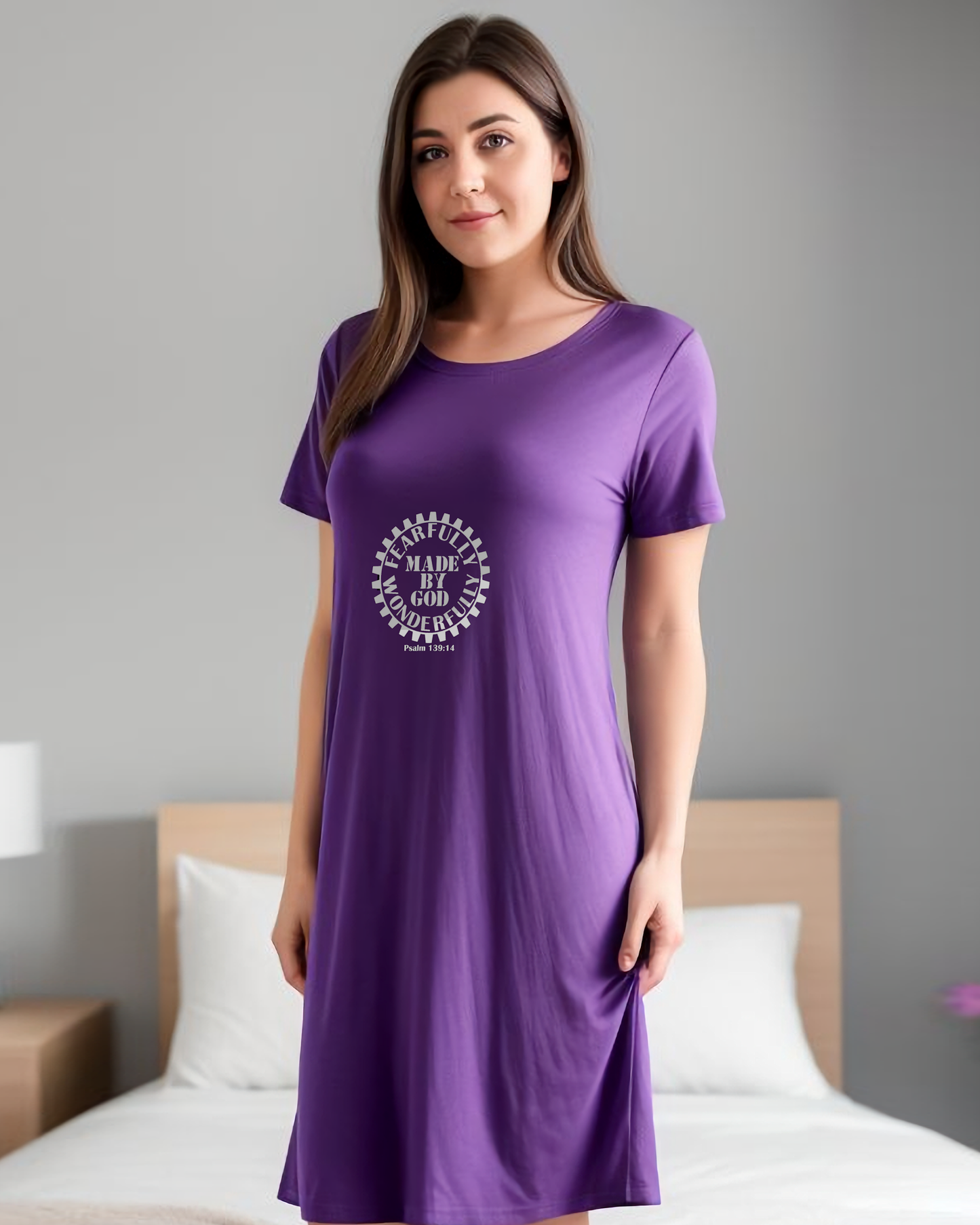 A woman in a bedroom wearing a purple t-shirt dress with a white Heavenly Dreamwear logo comprised of  text "Fearfully Wonderfully Made by God" inside a geared wheel.