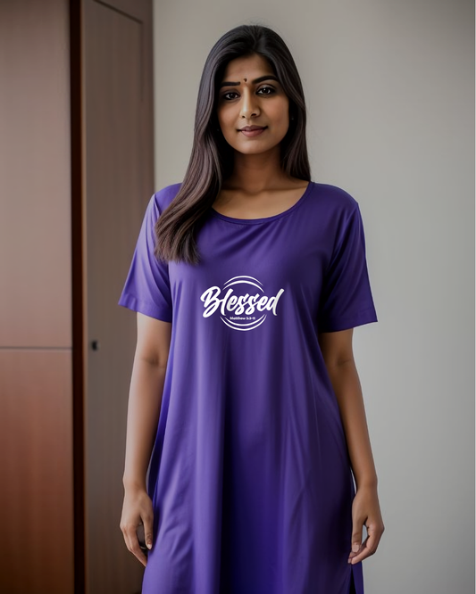 A woman in a bedroom wearing a purple Heavenly Dreamwear t-shirt dress with a white logo comprised of the text "Blessed" on top of a broken double circle with a biblical scripture reference beneath the text.