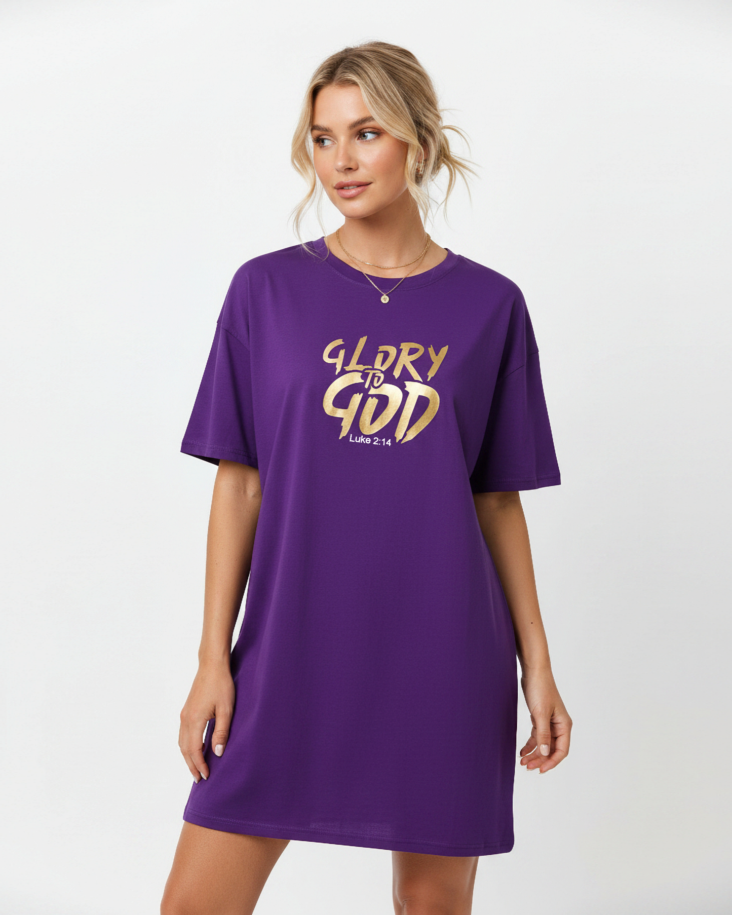 A woman modeling an oversized purple short-sleeve t-shirt dress with 'GLORY TO GOD' printed in gold across the chest, set against a plain light gray background.