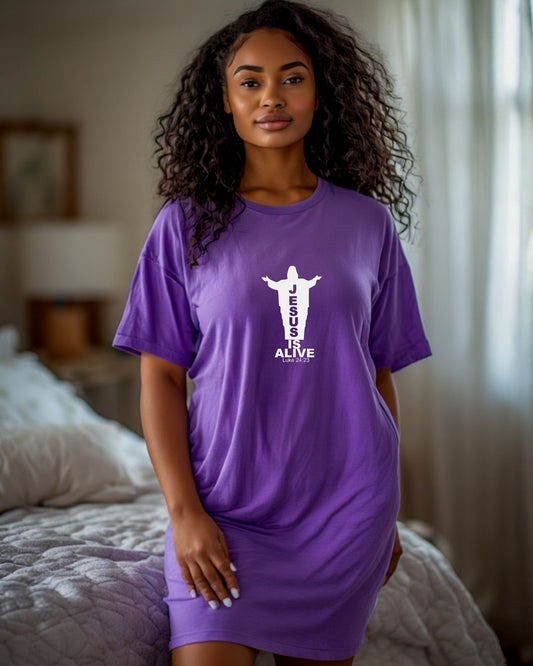 Woman in a bedroom wearing a purple t-shirt dress with a white Jesus silhouette graphic and text.