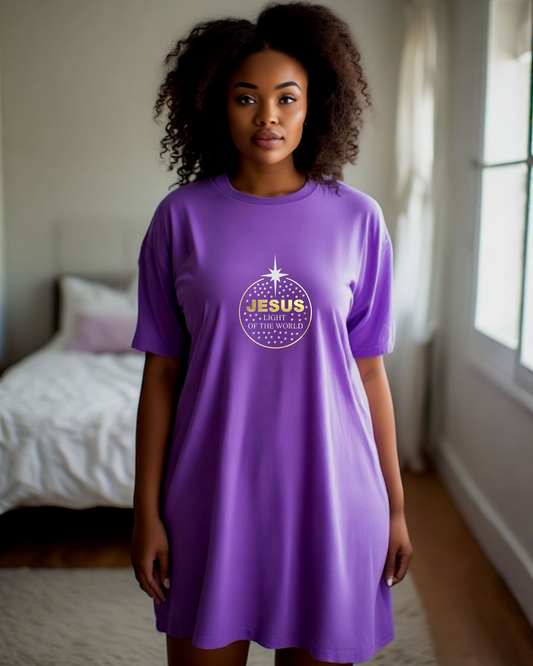 A woman wearing a purple short-sleeve t-shirt dress with a circular graphic on the chest that reads 'JESUS LIGHT OF THE WORLD' in gold and silver, standing in a bedroom.