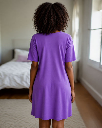 A woman with curly dark hair is shown from behind wearing a purple short-sleeve t-shirt dress in a bright bedroom.