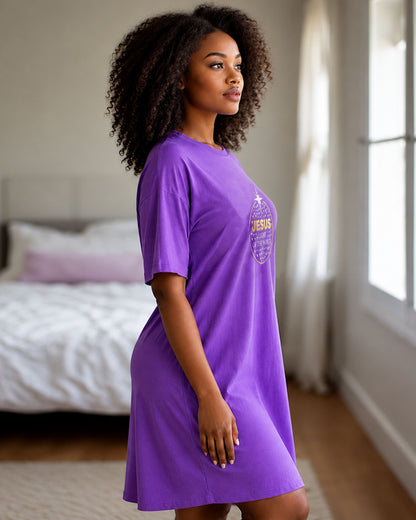A woman wearing a purple short-sleeve t-shirt dress with a gold and silver graphic on the chest, standing in a bedroom with a bed and window in the background.