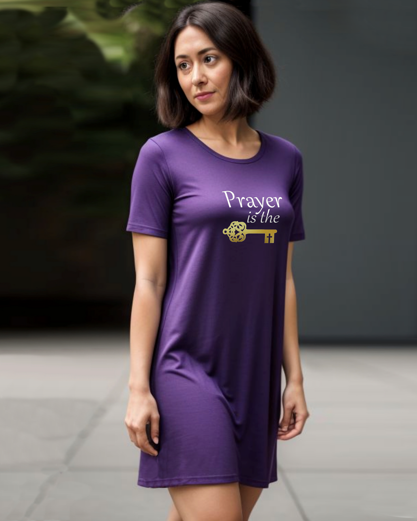 A woman is wearing a purple cotton t-shirt dress featuring a design by Heavenly Dreamwear.  The design includes white text that reads, "Prayer is the," followed by a gold key symbol.