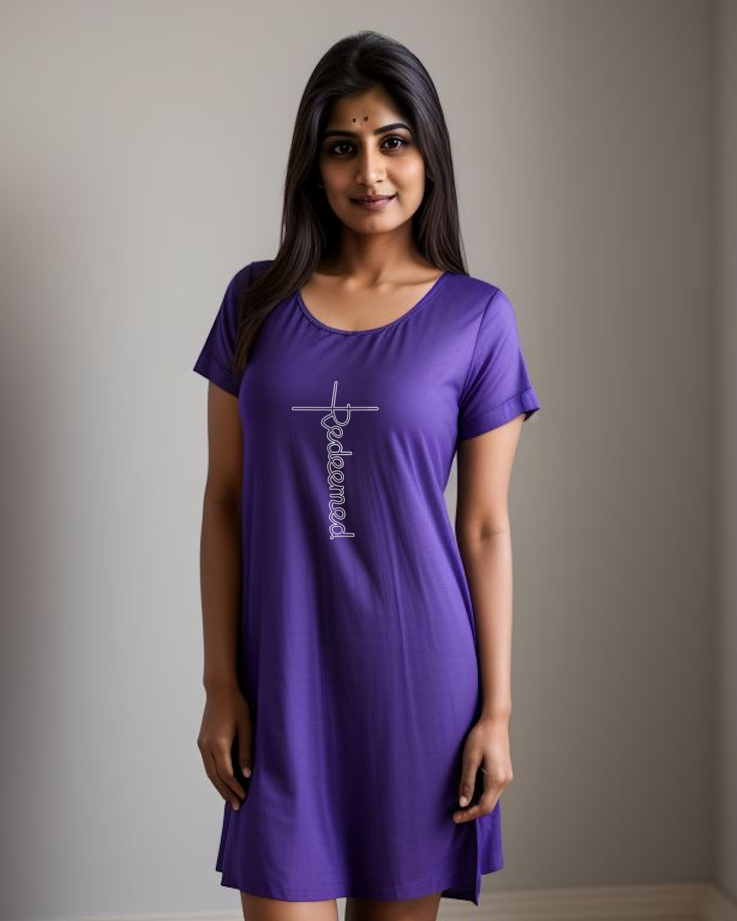 A woman in a bedroom wearing a purple T-shirt dress with an elaborate  white Heavenly Dreamwear designed 'Redeemed' logo printed on the front. 