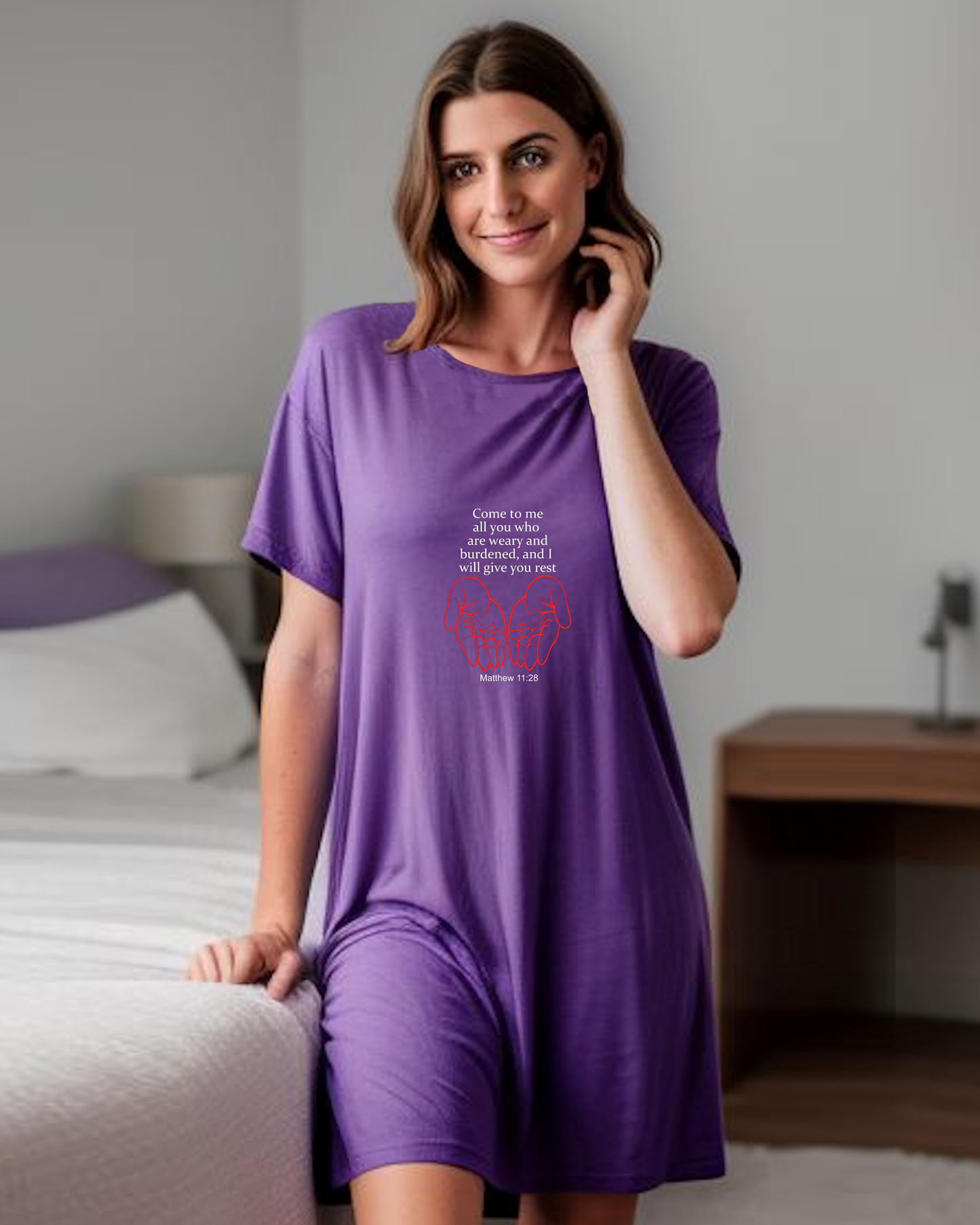 A woman in a bedroom wearing a purple Heavenly Dreamwear t-shirt dress with a white "come to me all you who are weary" text with two red hands facing palm up logo printed on the front.