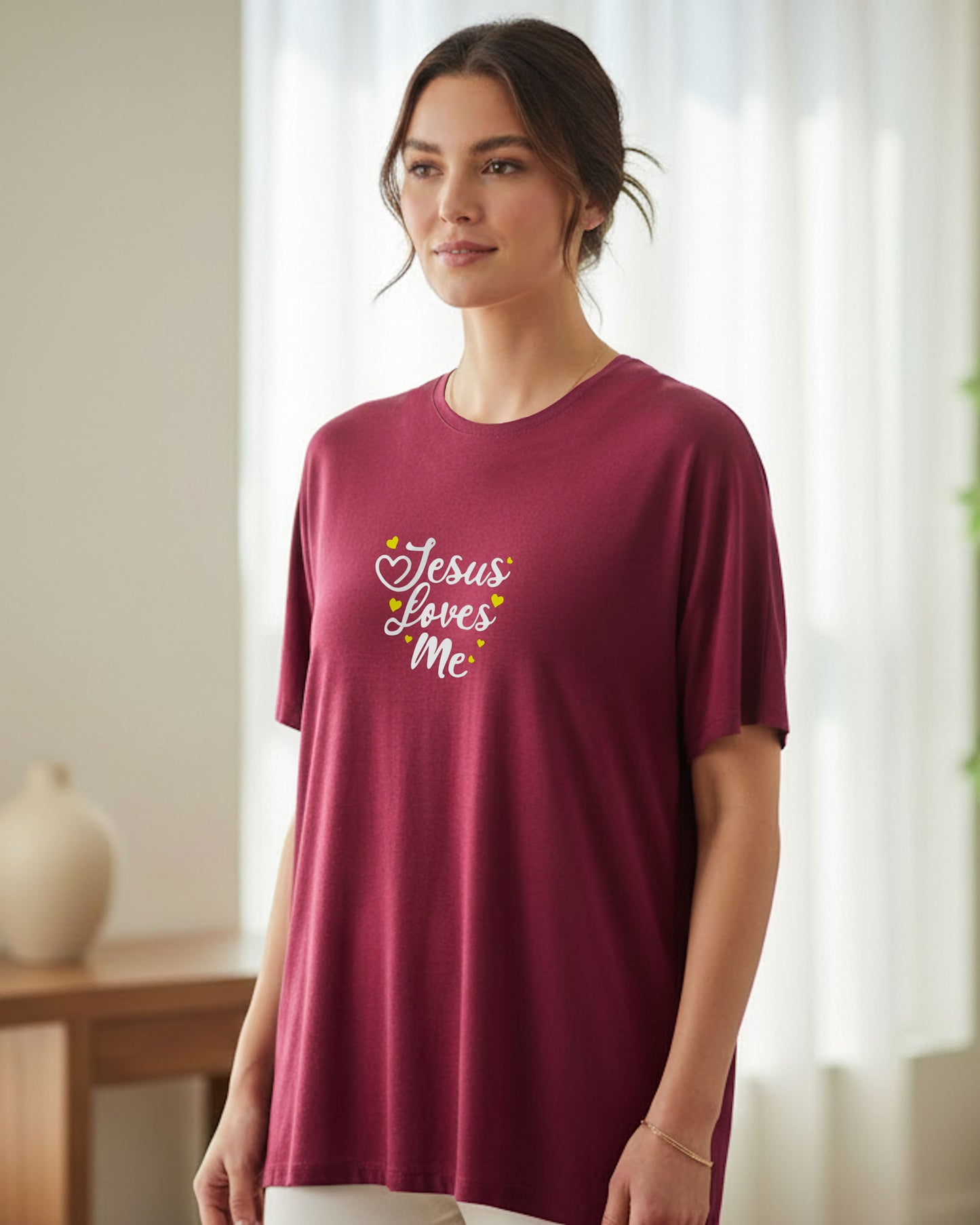 A woman indoors wearing a maroon t-shirt with white 'Jesus Loves Me' text and yellow hearts