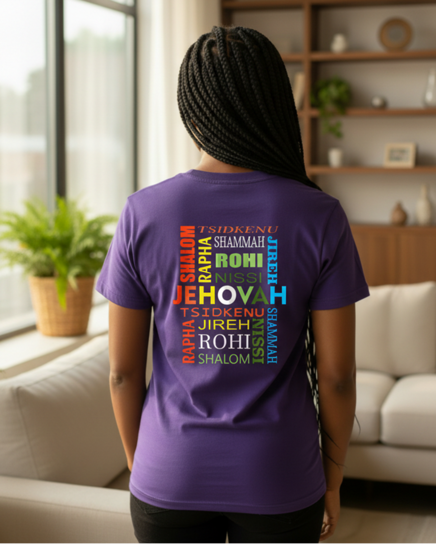 A woman viewed from behind wearing a purple short-sleeve t-shirt with a colorful word cloud graphic on the back. The graphic features various God's Hebrew names in red, orange, white, green, blue, and yellow.