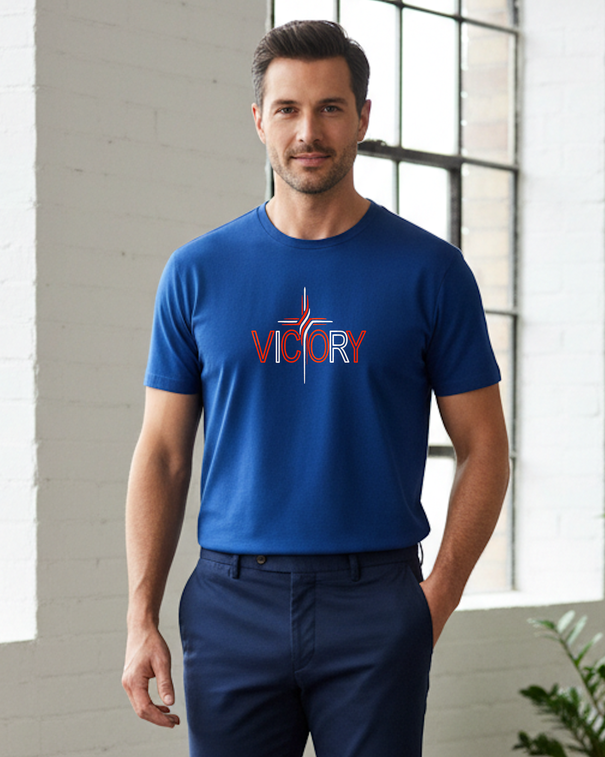 A man wearing a royal blue crewneck t-shirt with a red and white 'VICTORY' graphic on the chest, paired with navy blue trousers, standing in a bright, modern room with white brick walls and a large window.