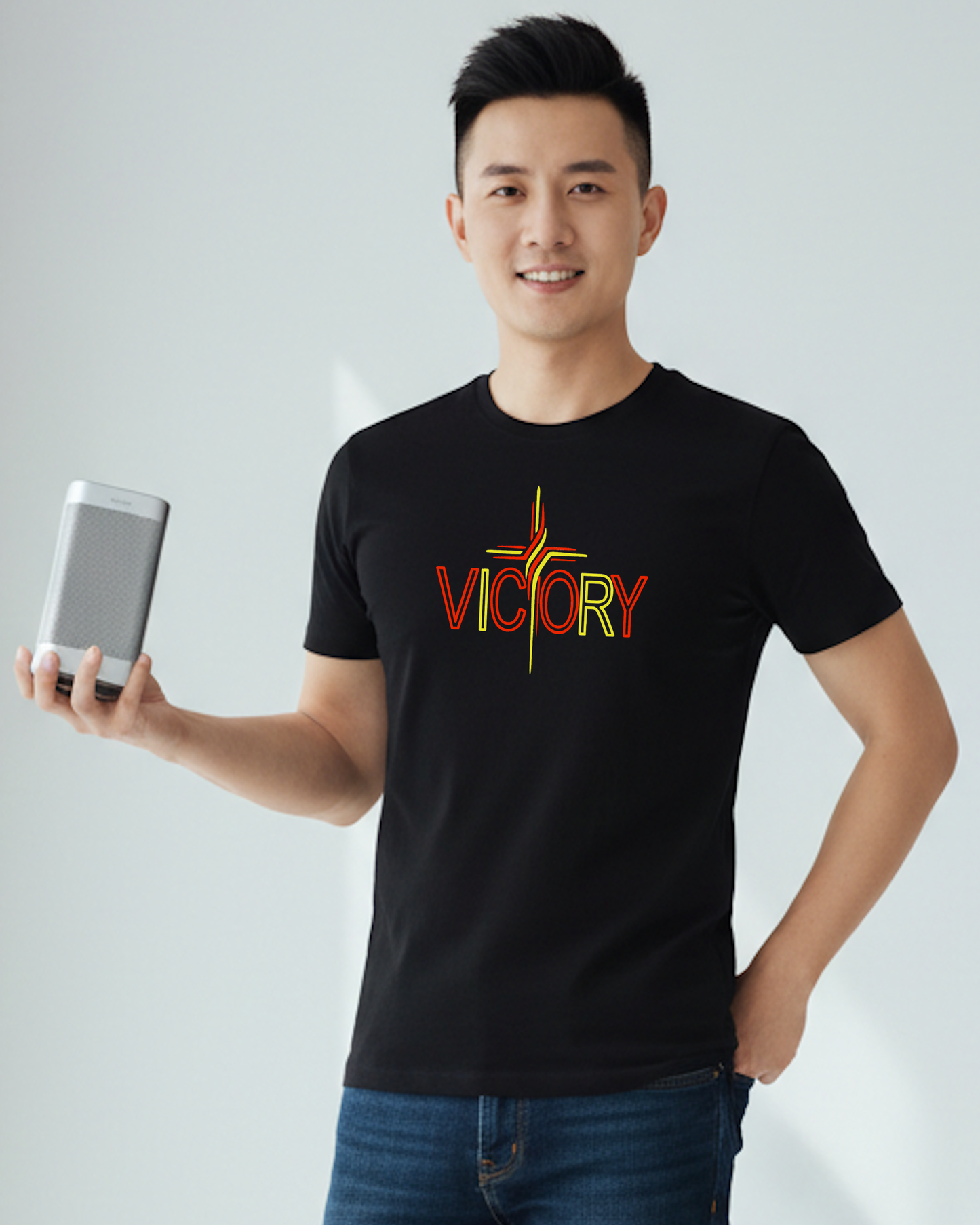 A smiling man wearing a black t-shirt with a colorful 'VICTORY' graphic on the chest, holding a speaker in his right hand.