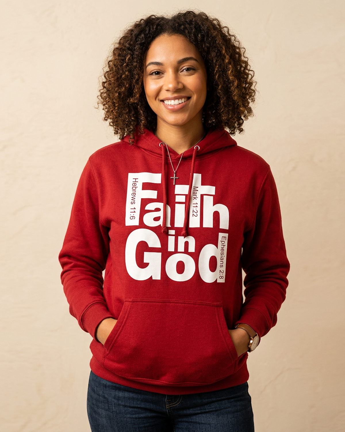 A smiling woman with curly hair wearing a red hoodie with a white graphic that reads 'Faith in God' and Bible verse references, paired with dark blue jeans, standing against a textured beige wall.