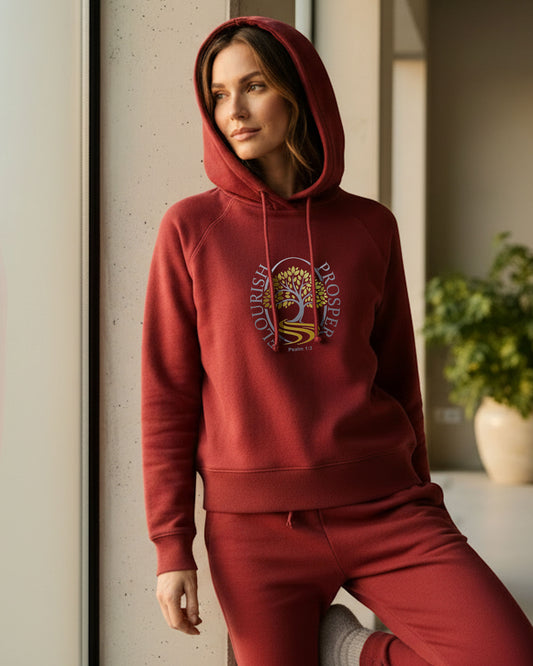 Woman standing near a window wearing a red hoodie with a gold/silver logo  with text "Flourish Prosper" around a tree.