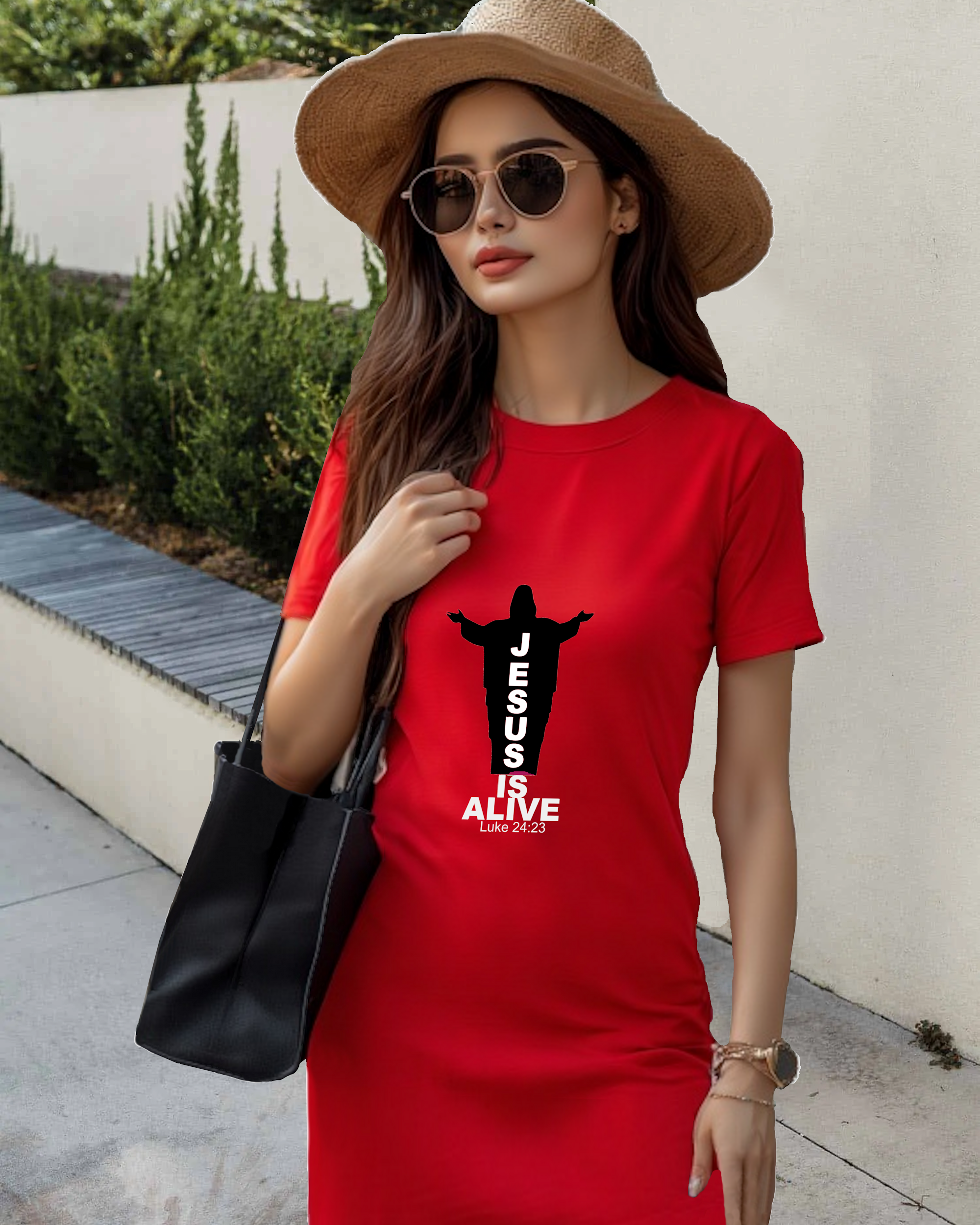 A woman in a garden wearing a red Heavenly Dreamwear t-shirt dress with a black and white logo of a silhouette of Jesus with the name "JESUS" embedded in it. Underneath is the text "IS ALIVE" with the scriptural reference: Luke 24:23.