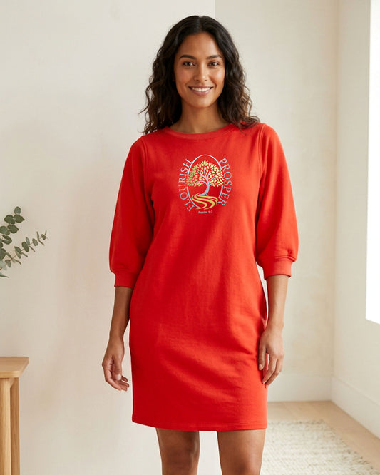 A woman wearing a bright red crewneck dress with three-quarter sleeves and a gold and silver 'Flourish Prosper' tree graphic on the chest, standing in a brightly lit indoor room.
