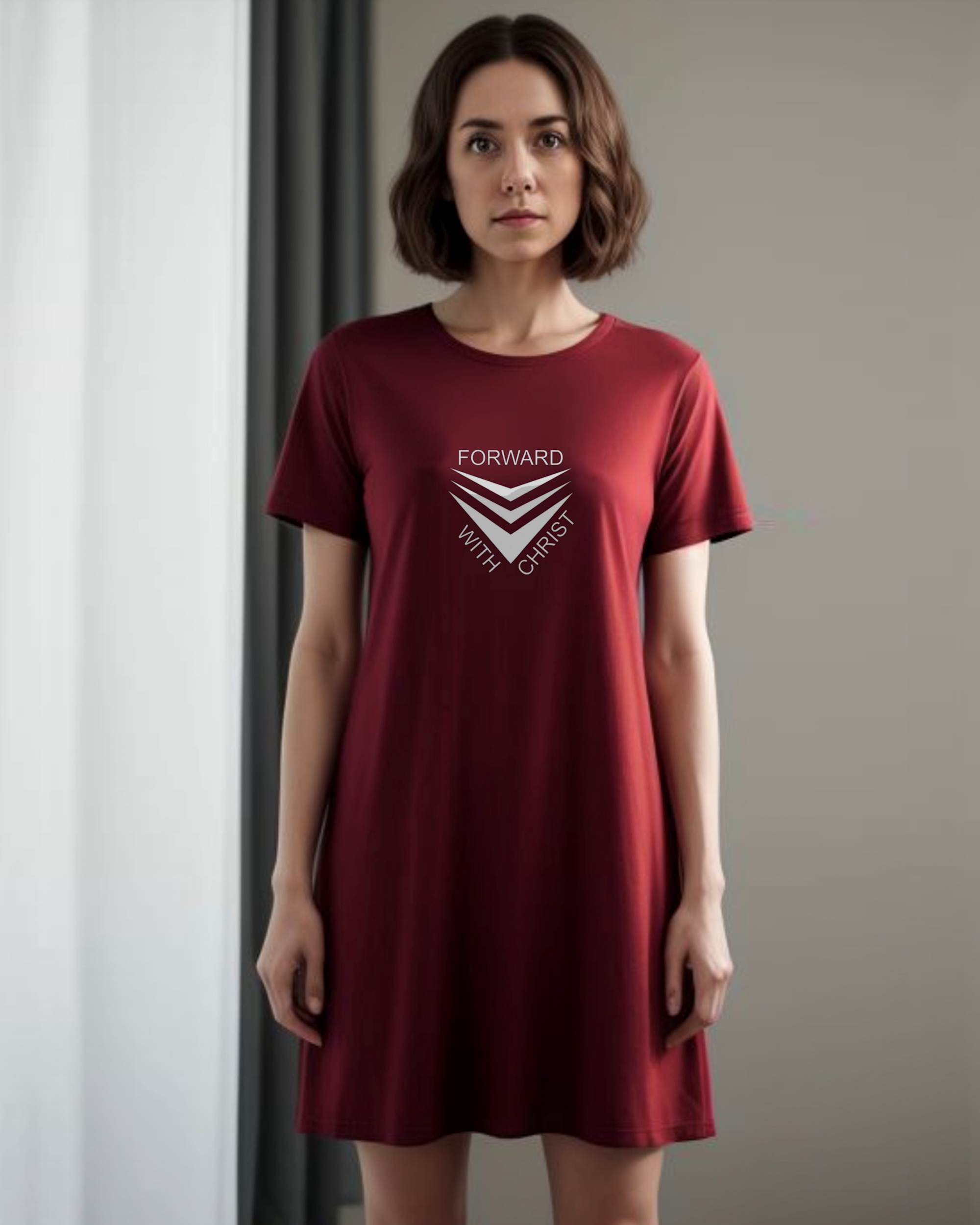 A woman standing in a lounge wearing a red Heavenly Dreamwear t-shirt dress with a white logo made up of  3 chevrons surrounded by the text, "Forward With Christ".