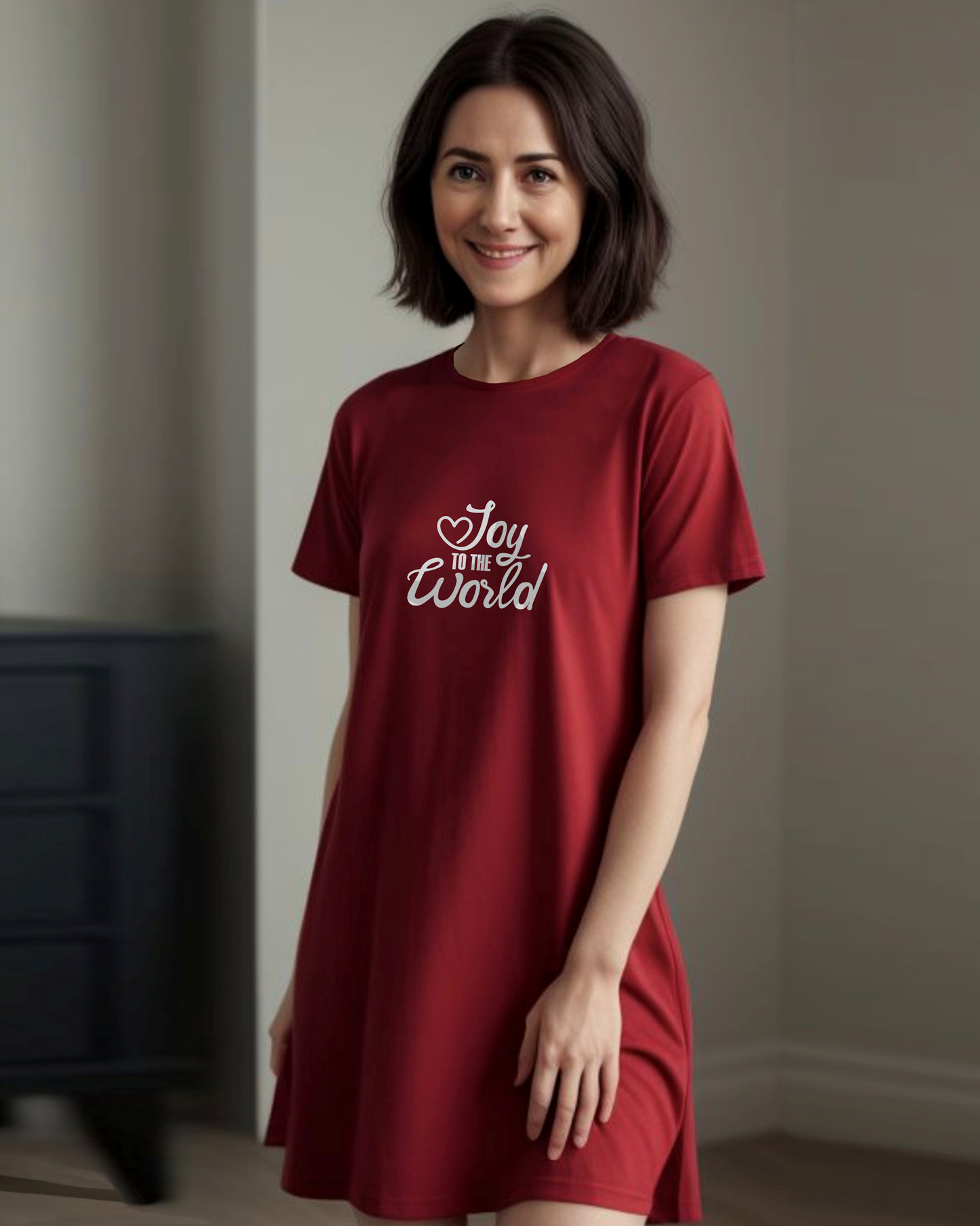 A woman standing indoors wearing a red Heavenly Dreamwear t-shirt dress with a white "Joy to the World" logo printed on the front.