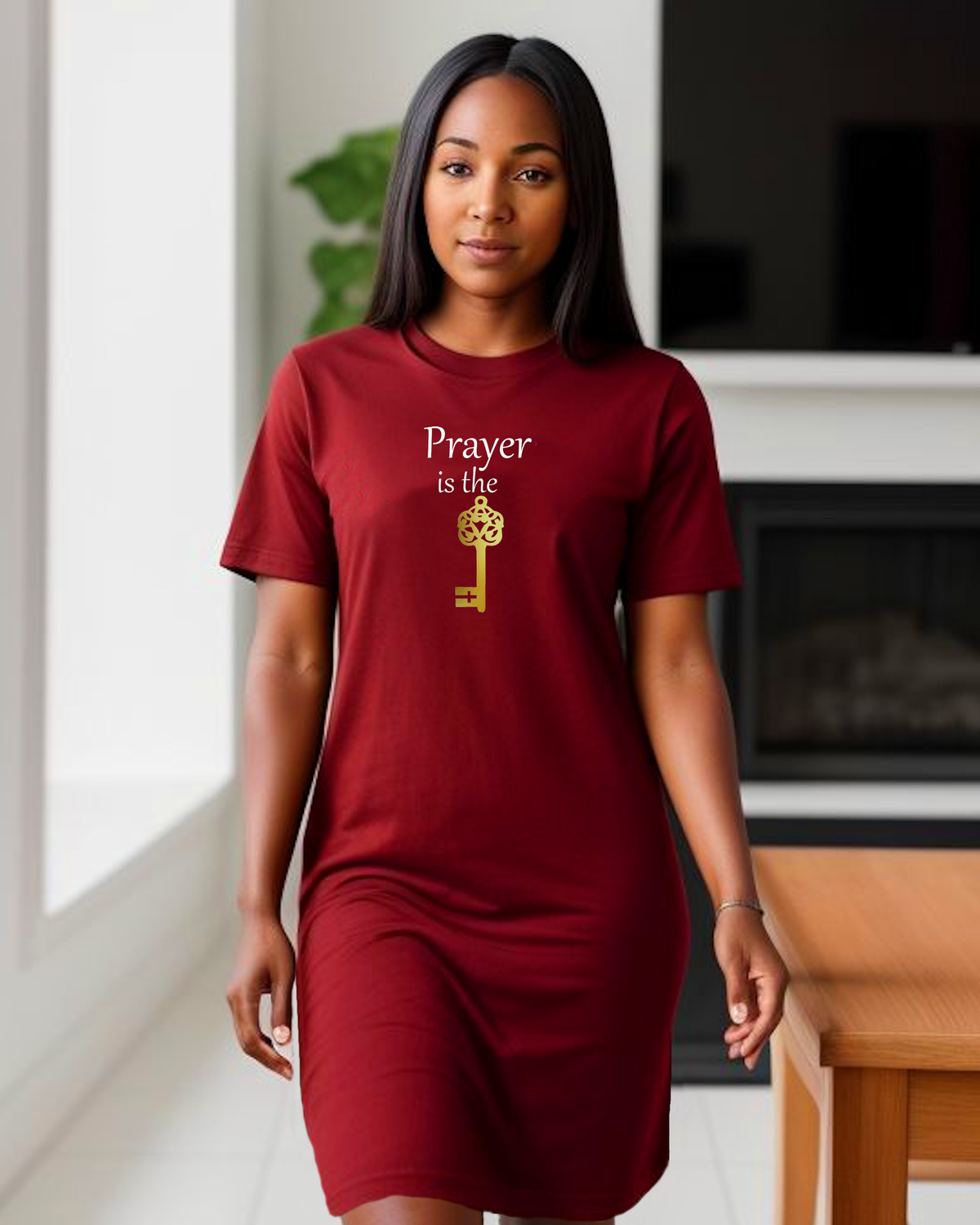 A woman wearing a red t-shirt dress with a graphic that reads 'Prayer is the key' in white and gold, standing in a modern living room setting.