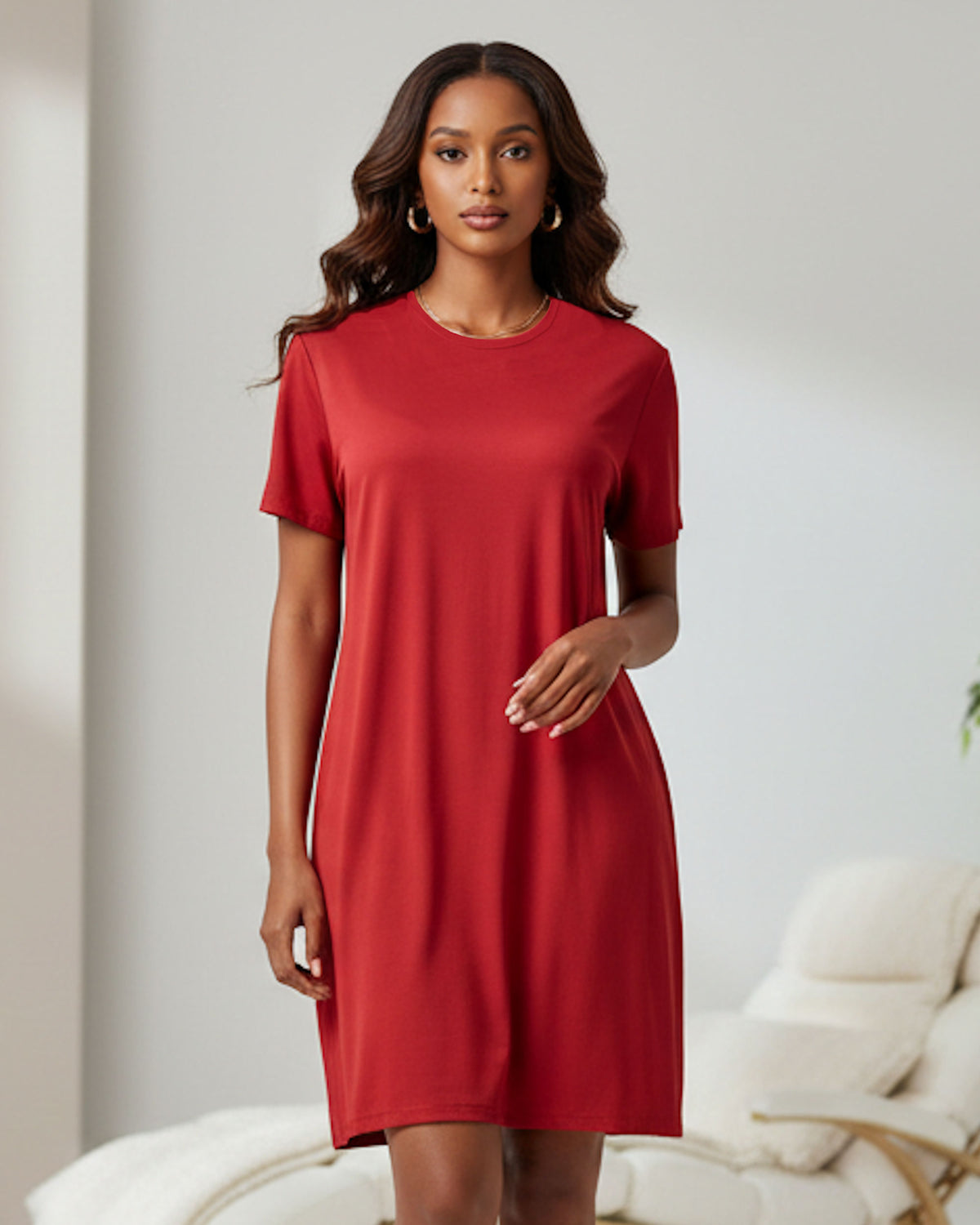 A woman modeling a short-sleeve red t-shirt dress in a brightly lit, modern room with a white sofa in the background.