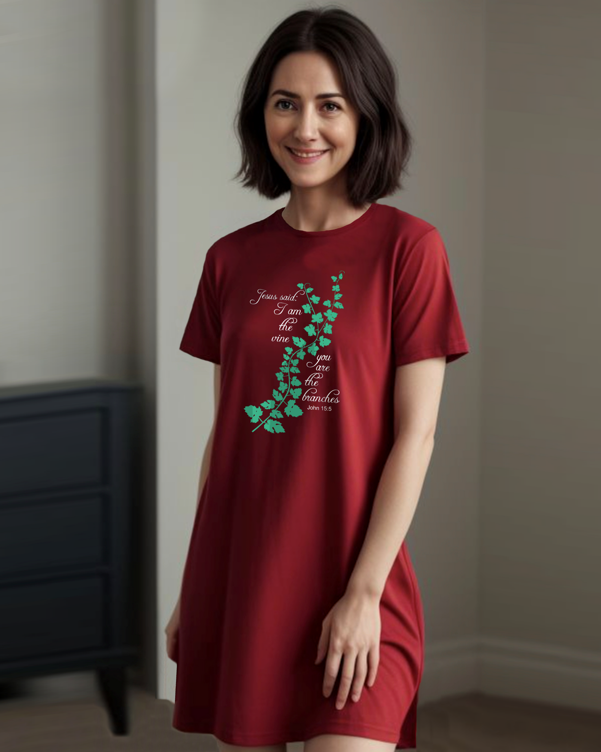 Woman standing in a bedroom wearing a red t-shirt dress with a logo made up of a green vine draped across the front with a biblical scripture on either side in white text from John 15:5