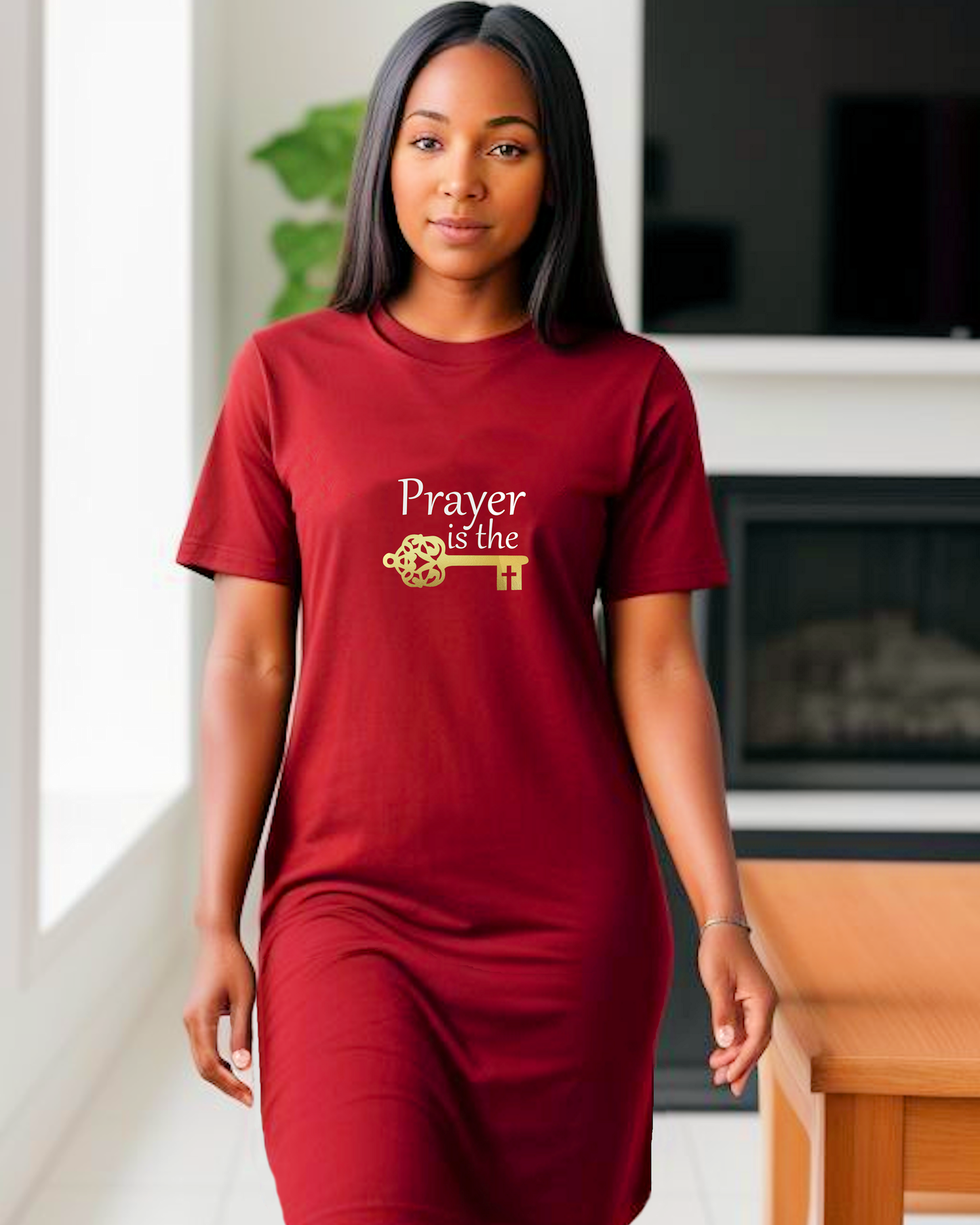 A woman standing in a dining room wearing a red t-shirt dress featuring a logo design by Heavenly Dreamwear.  The logo includes white text that reads, "Prayer is the," followed by a gold key symbol.
