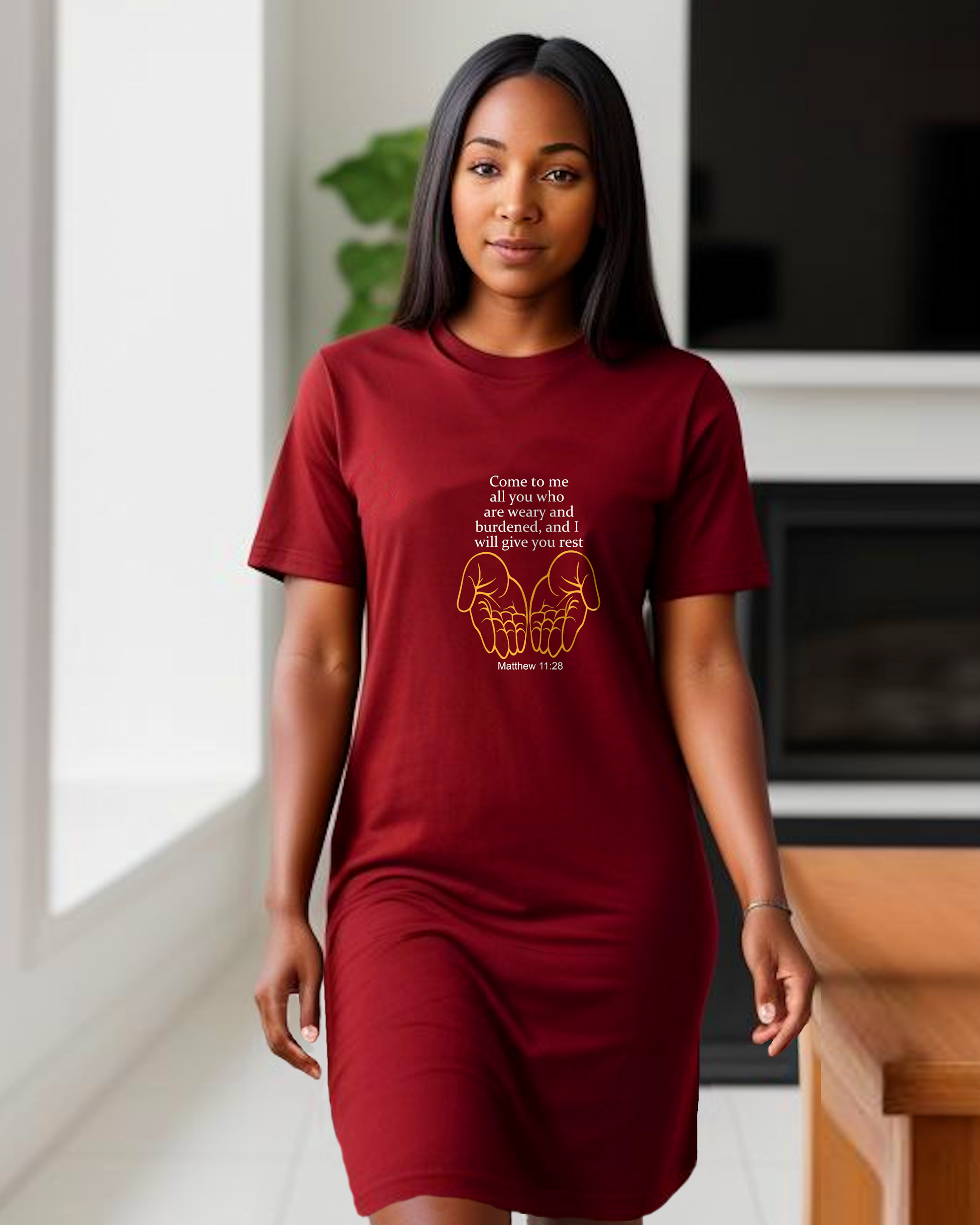 A woman standing in a kitchen wearing a red Heavenly Dreamwear t-shirt dress with white "come to me all you who are weary" text with two yellow hands facing palm up logo printed on the front.