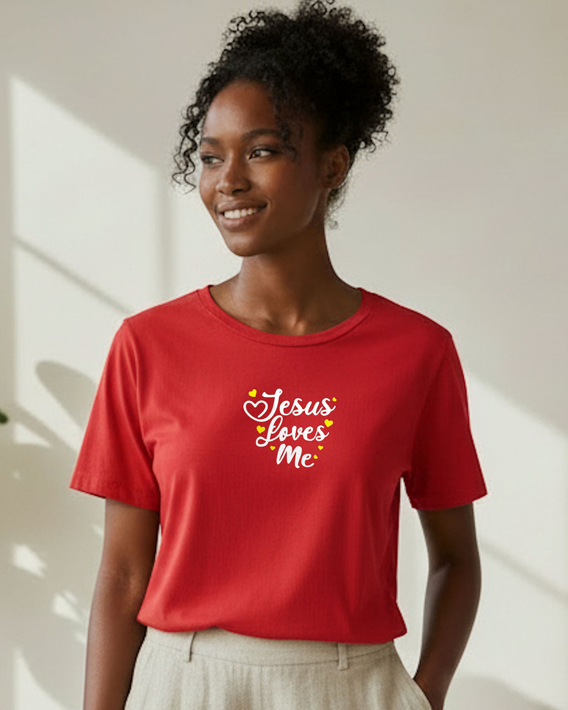 A smiling woman standing in a white room wearing a red t-shirt with white text, 'Jesus loves me', with yellow hearts.