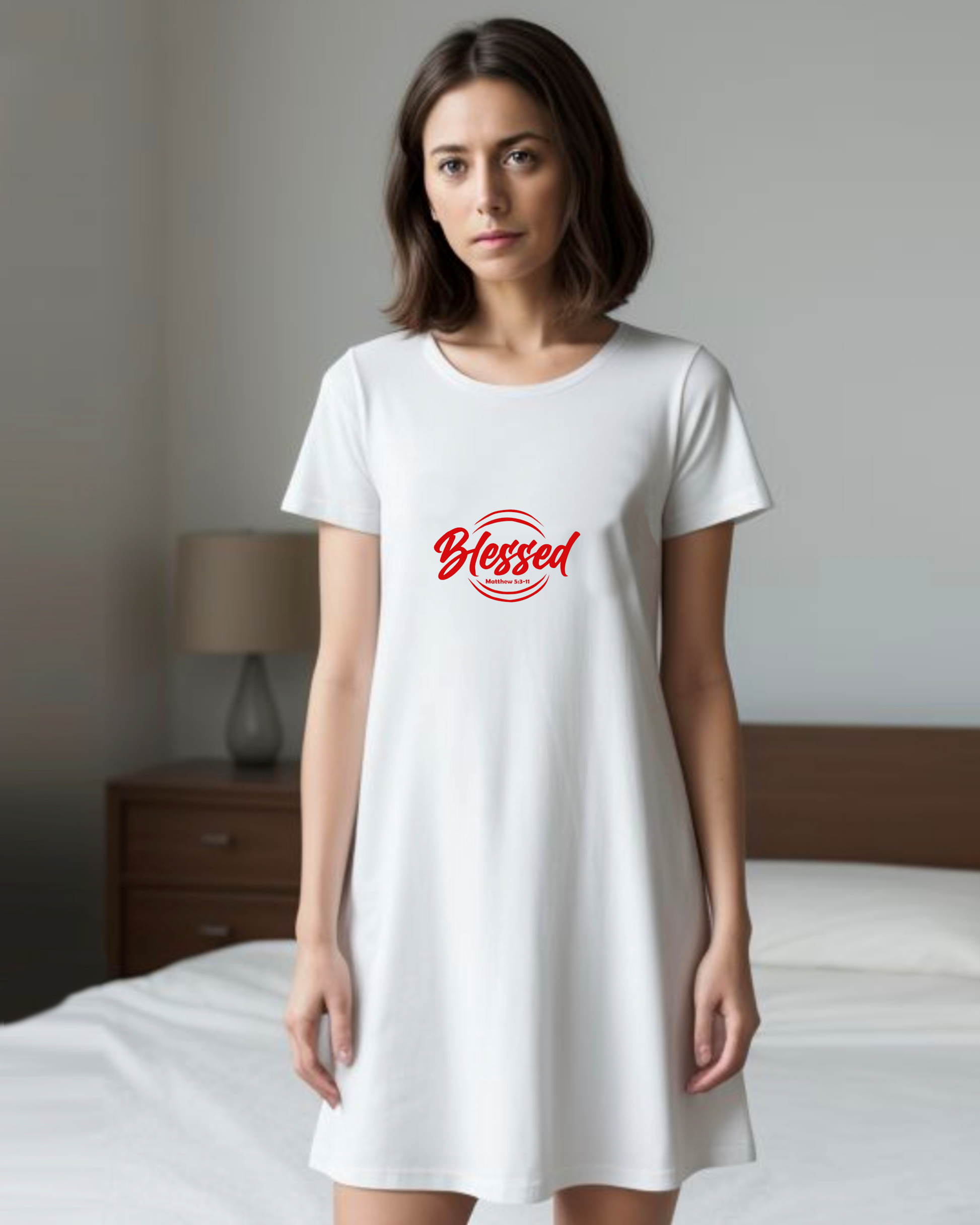 A woman in a bedroom wearing a white Heavenly Dreamwear t-shirt dress with a red logo comprised of the text "Blessed" on top of a broken double circle with a biblical scripture reference beneath the text.