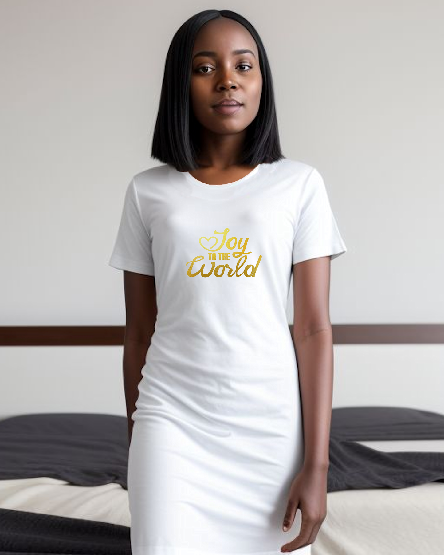 A woman standing in a bedroom wearing a white Heavenly Dreamwear t-shirt dress with a gold "Joy to the World" logo printed on the front.
