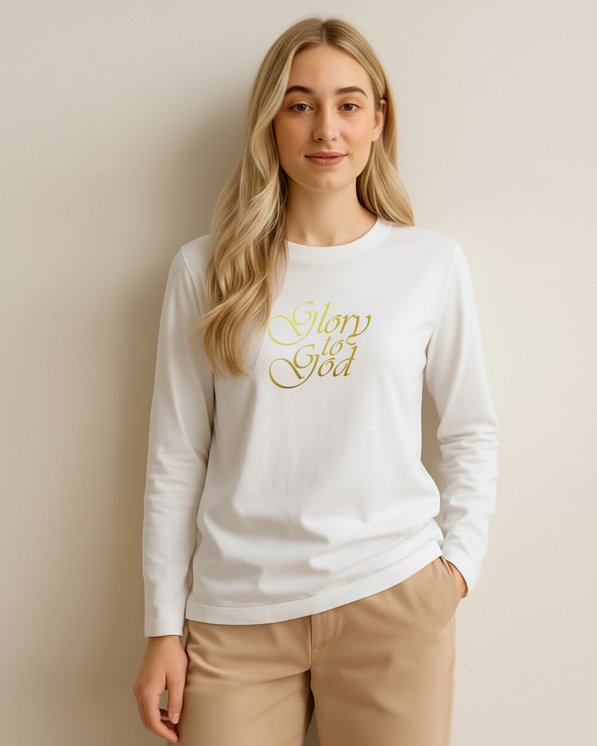 A woman modeling a white long-sleeve crewneck t-shirt with 'Glory to God' printed in gold cursive across the chest, paired with beige pants against a plain cream white background.