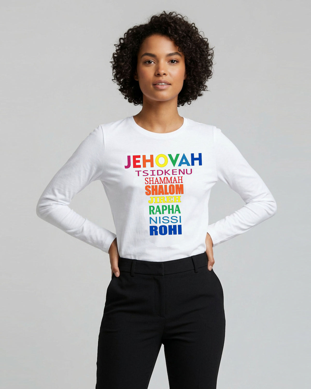A woman modeling a white long-sleeve t-shirt with a colorful rainbow text graphic on the chest, paired with black pants, against a solid gray studio background.