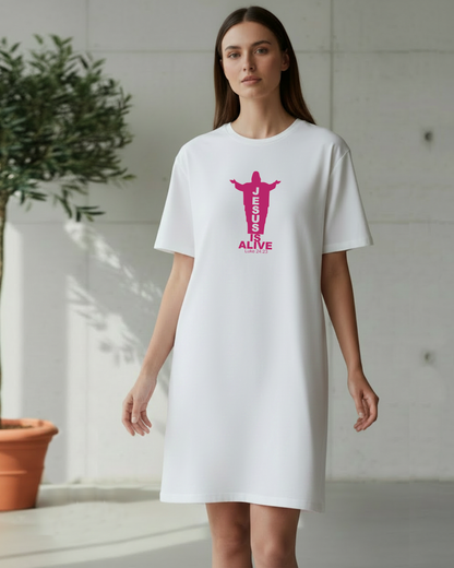 A woman modeling a white short-sleeve t-shirt dress with a pink graphic of Jesus Christ and the text 'JESUS IS ALIVE' on the front, set against a minimalist concrete wall background with a potted plant.
