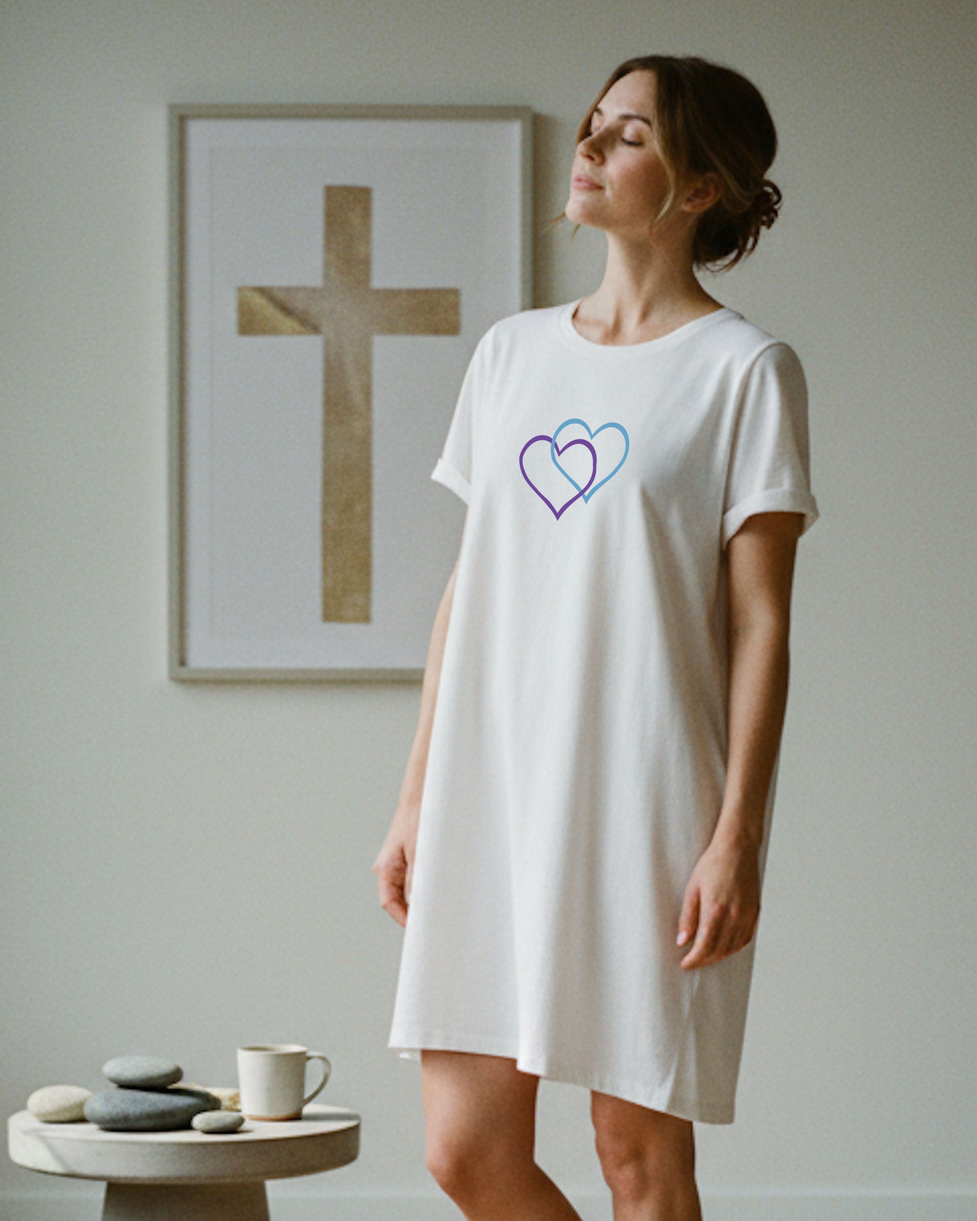 A woman standing indoors wearing a white t-shirt dress with a Heavenly Dreamwear logo made of an purple and blue intertwined heart shapes printed on the front.