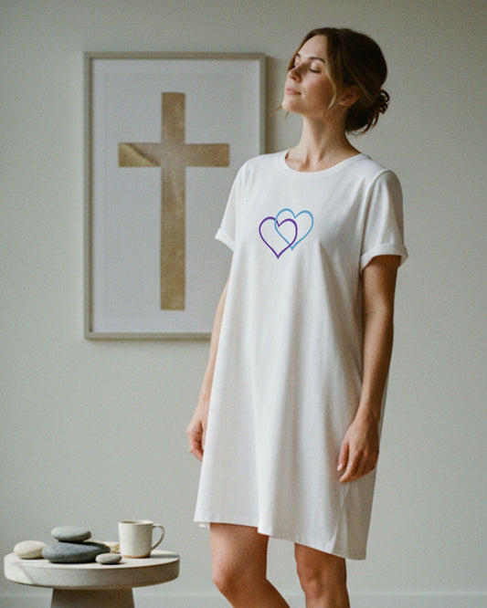 A woman standing indoors wearing a white t-shirt dress with a Heavenly Dreamwear logo made of an purple and blue intertwined heart shapes printed on the front.