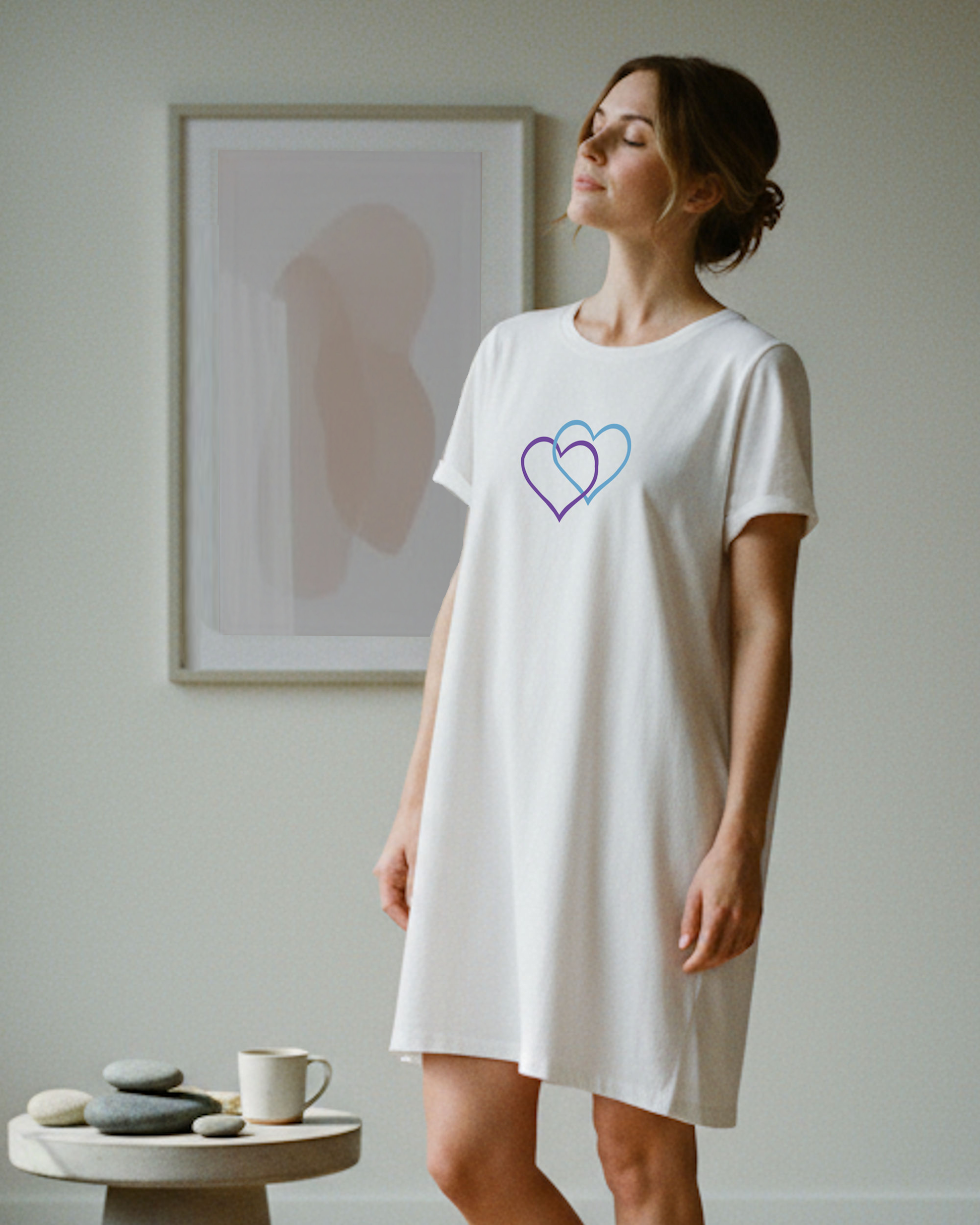 A woman standing indoors wearing a white t-shirt dress with a Heavenly Dreamwear logo made of an purple and blue intertwined heart shapes printed on the front.