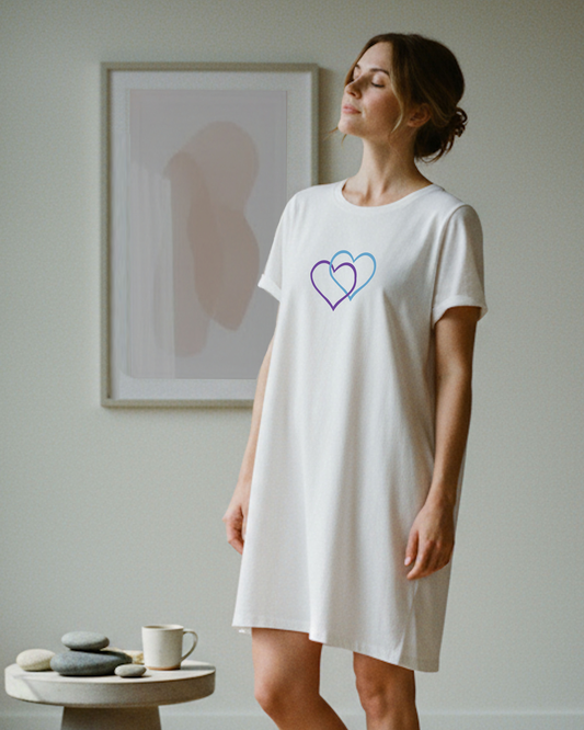 A woman standing indoors wearing a white t-shirt dress with a Heavenly Dreamwear logo made of an purple and blue intertwined heart shapes printed on the front.