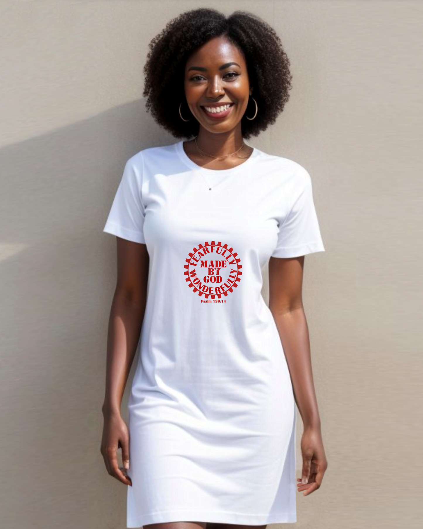 A woman standing against a wall wearing a white t-shirt dress with a red Heavenly Dreamwear logo comprised of  text "Fearfully Wonderfully Made by God" inside a geared wheel.