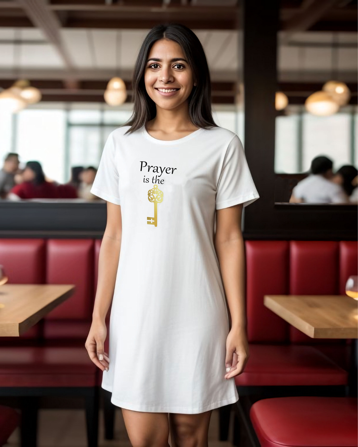 A woman wearing a white short-sleeve t-shirt dress with a graphic print that reads 'Prayer is the key' in black text and a gold key design, standing in a restaurant with red booths in the background.
