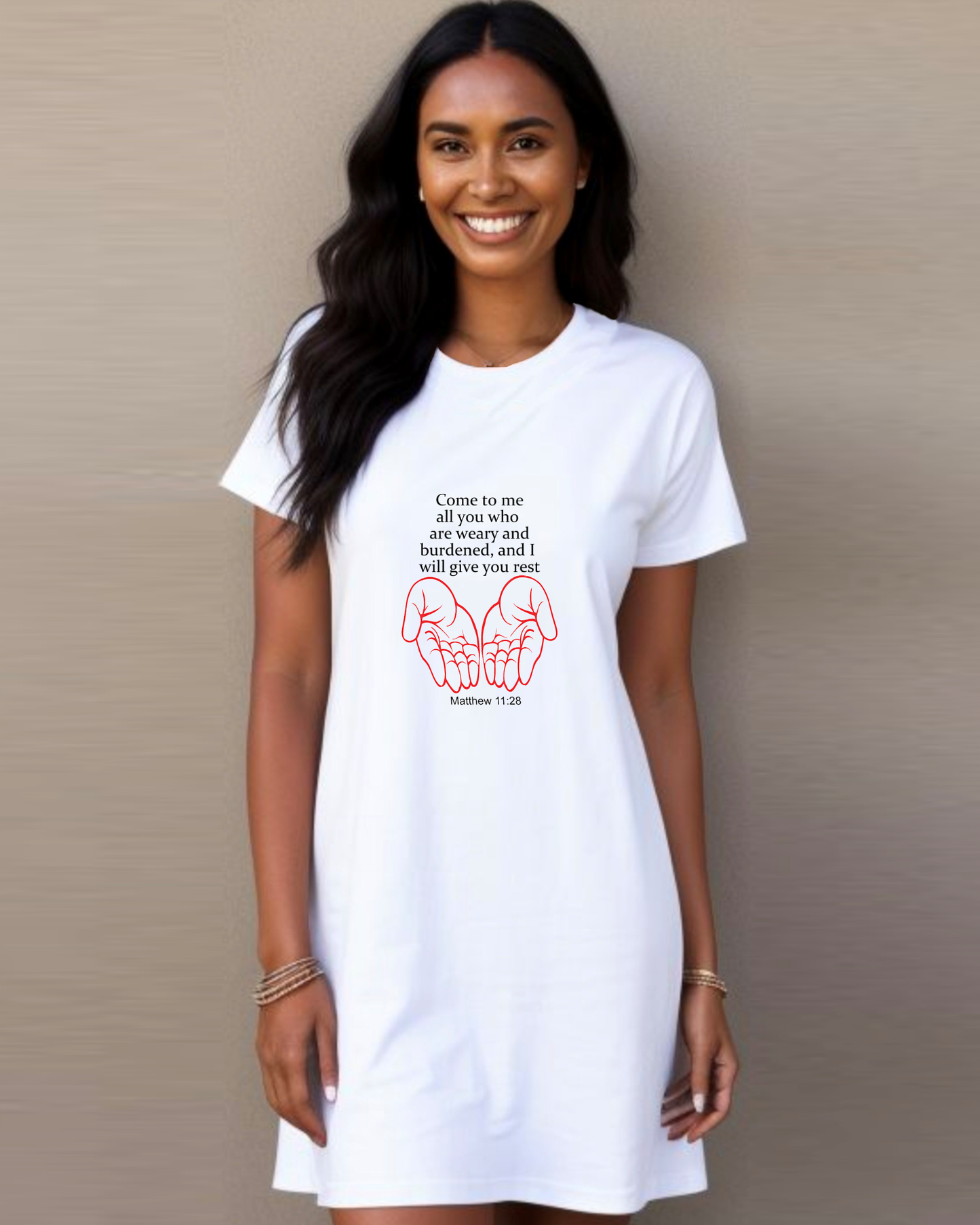 A woman wearing a white Heavenly Dreamwear t-shirt dress with black "come to me all you who are weary" text with two red hands facing palm up logo standing against a beige wall.